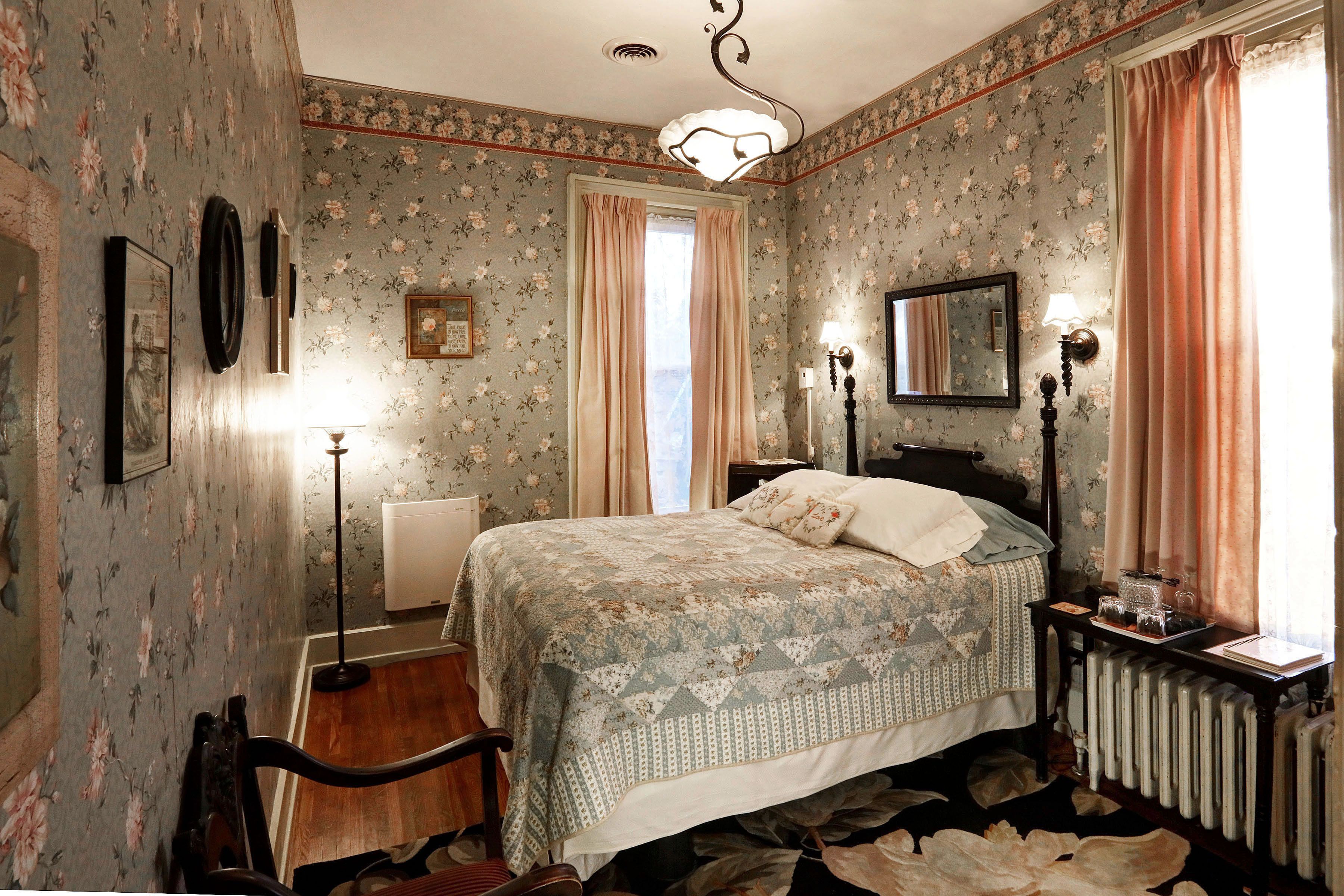 A bedroom with floral wallpaper, a bed with a patterned bedspread, a nightstand with a lamp, and a radiator.