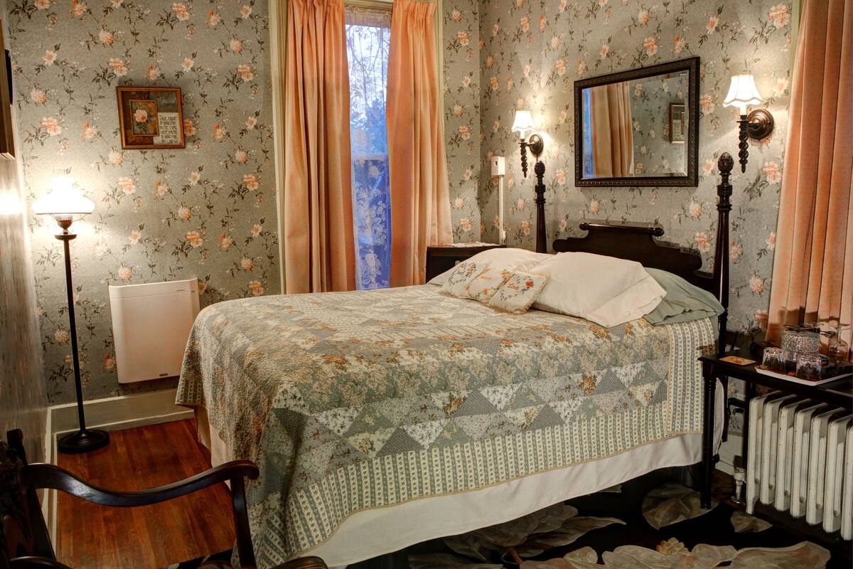 A bedroom with floral wallpaper, a bed with a patterned bedspread, two bedside lamps, a nightstand with a lamp, a radiator, and a chair.