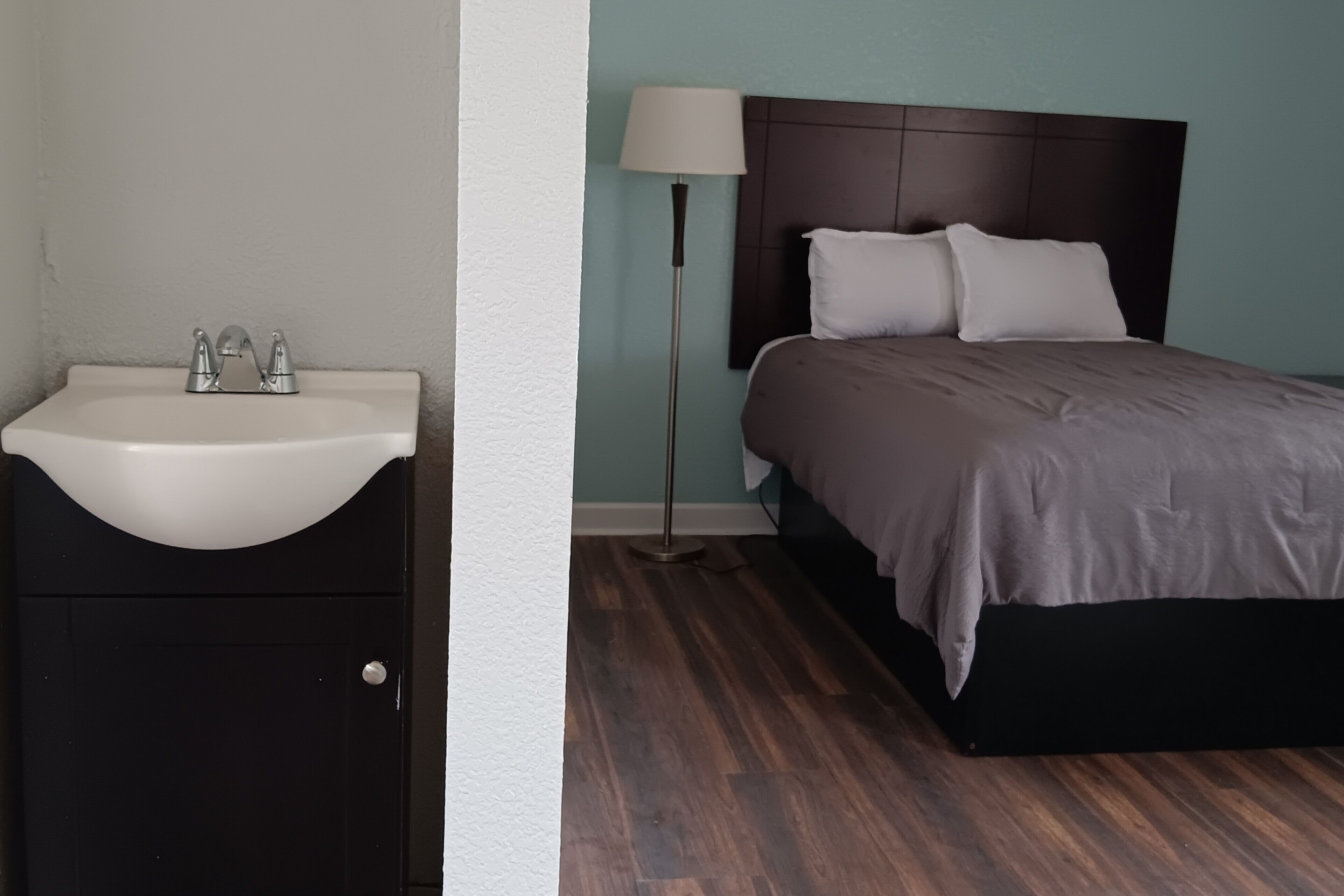 Un dormitorio con piso de madera, una cama con cabecera oscura, una lámpara de noche y un lavamanos blanco.