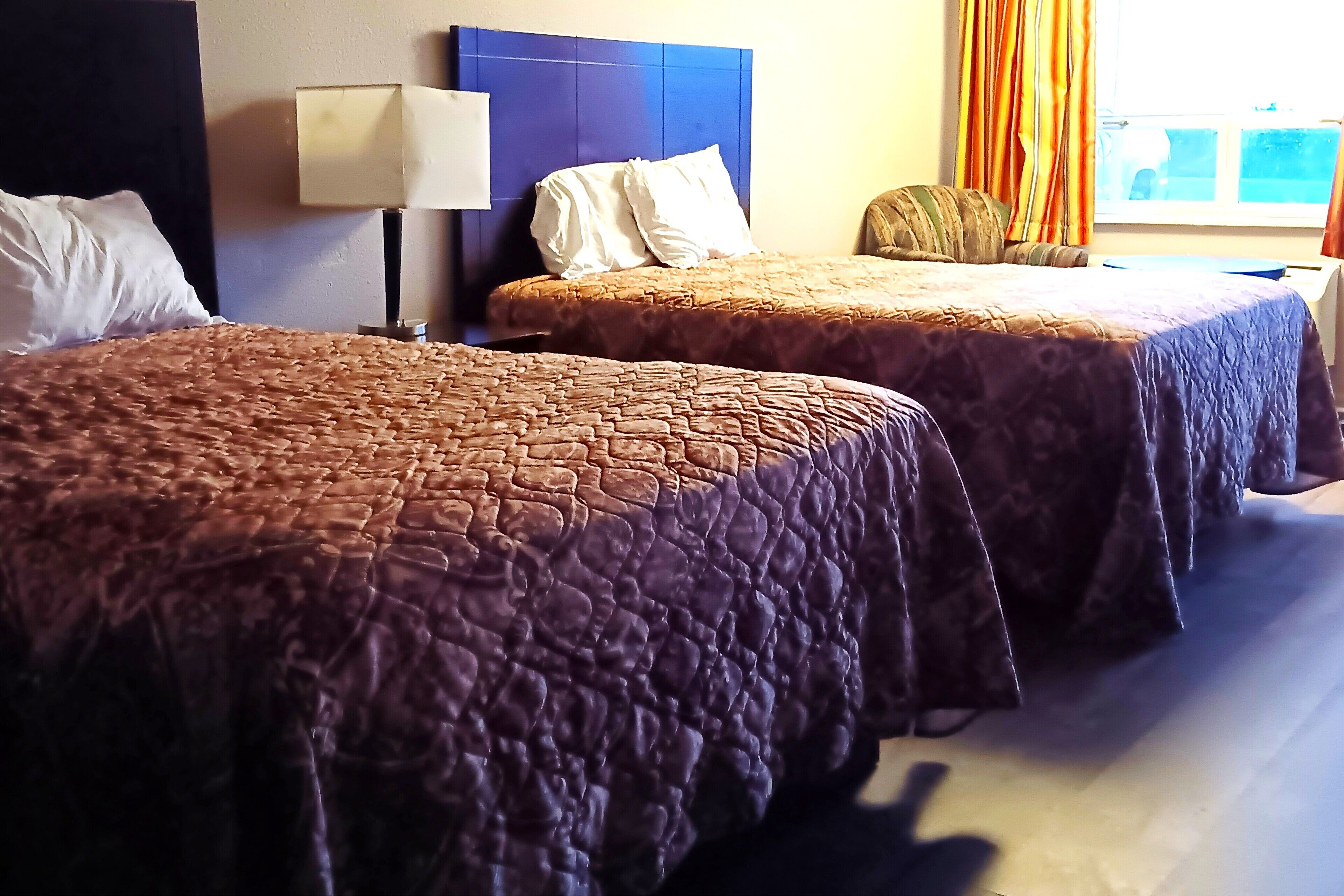 Two beds with brown quilts, a bedside lamp, and a window with curtains.