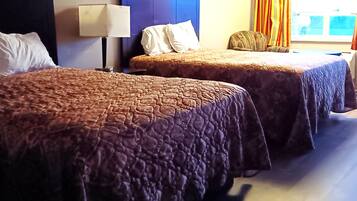 Two beds with brown quilts, a bedside lamp, and a window with curtains.