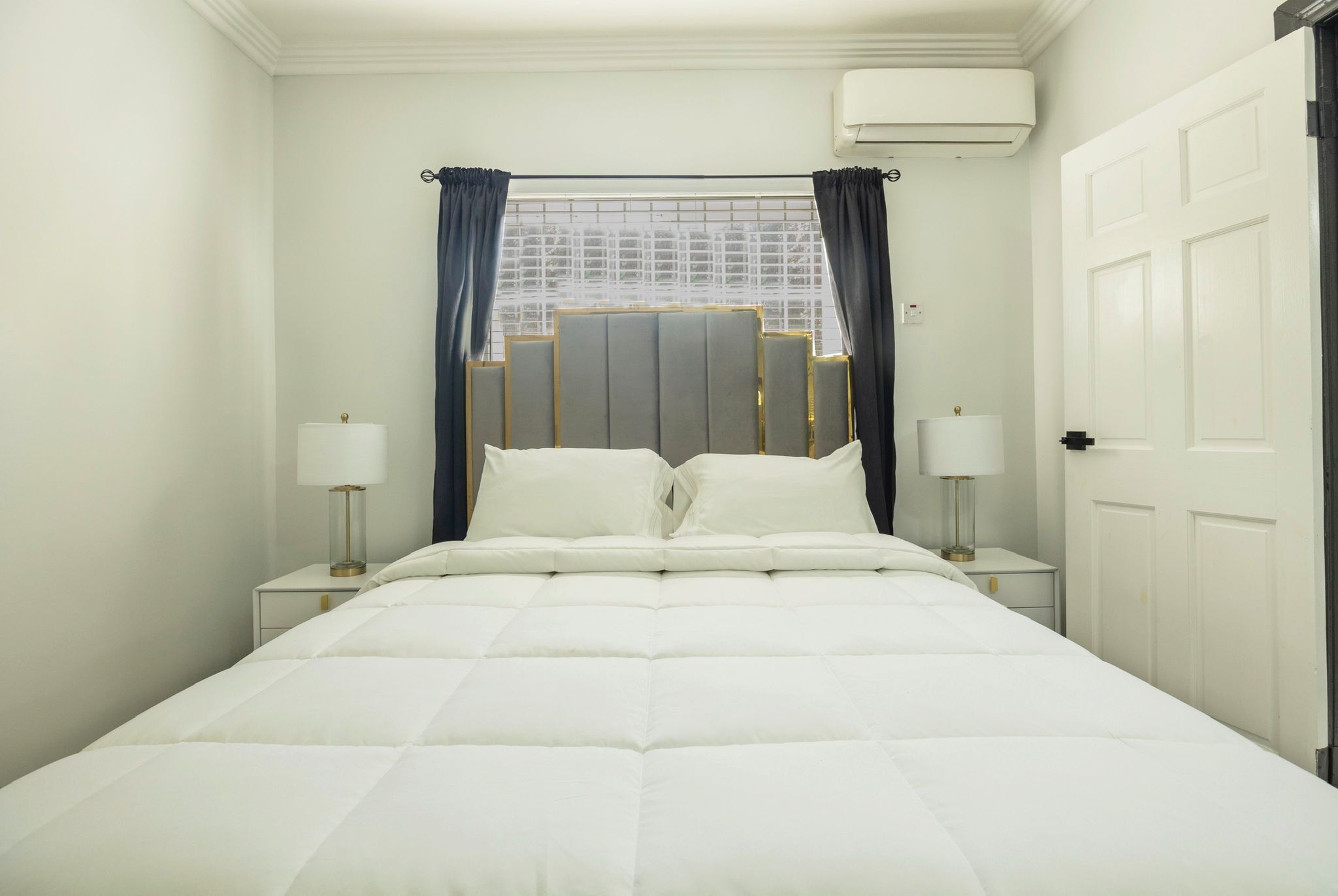 A neatly made bed with white bedding, two bedside lamps, and a window with curtains.