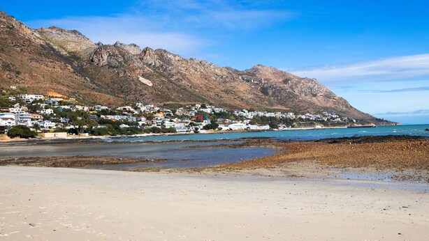 A coastal town with buildings on a hillside, a beach, and a rocky shoreline. - Anchor Bay (Cape Town)