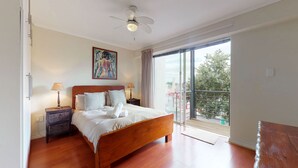 A bedroom with a wooden bed, two bedside tables with lamps, a ceiling fan, a large window with a view of the outdoors, and a painting on the wall. - Anchor Bay (Cape Town)