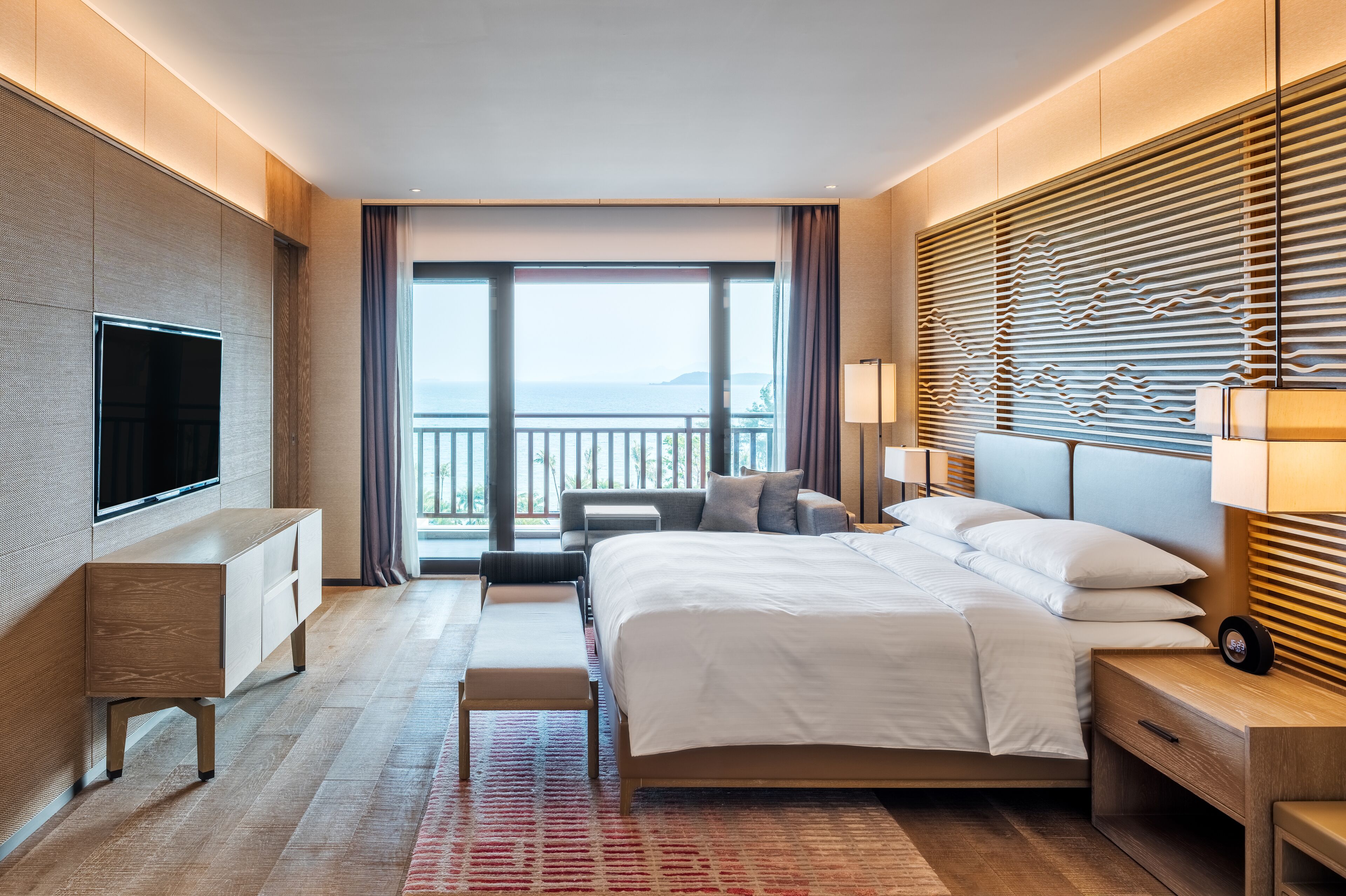 A modern hotel room with a large bed, a flat-screen TV, a bedside table, a bench, and a view of the sea through large windows.