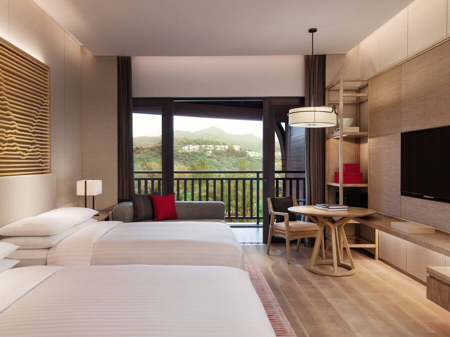A hotel room with a large bed, a desk, a chair, a TV, and a balcony with a view of the mountains.