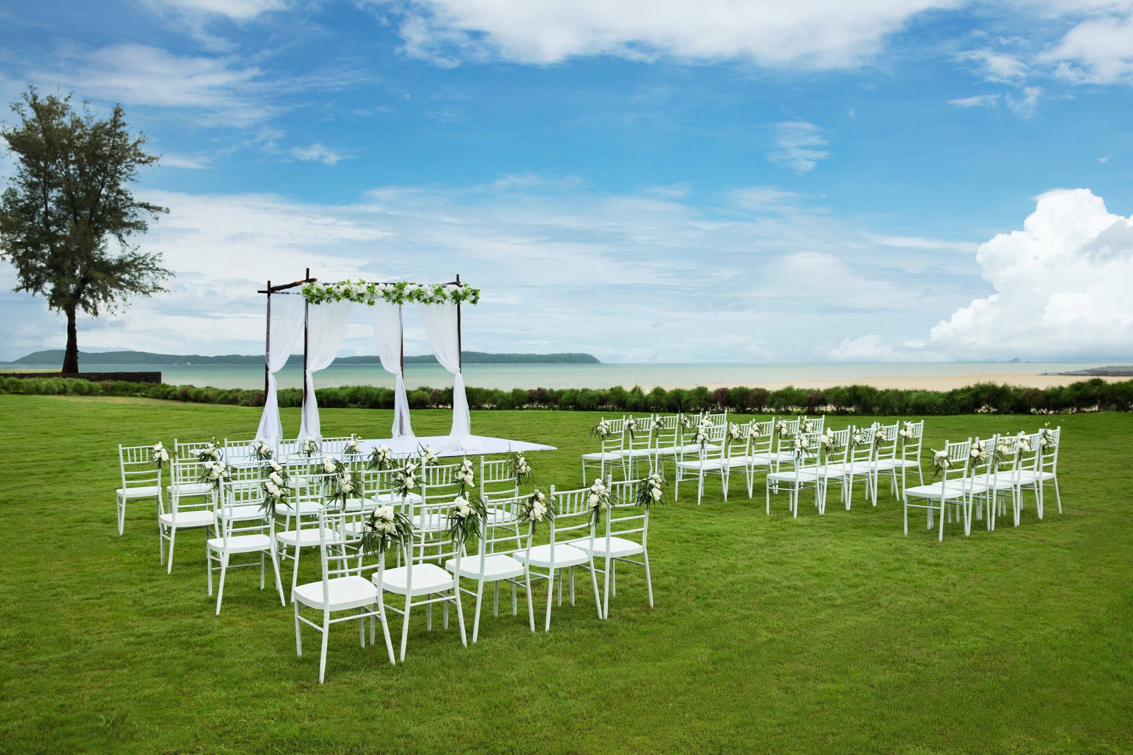 Montaje para una ceremonia de boda con sillas blancas y un dosel en un campo cubierto de hierba, con vista al mar.