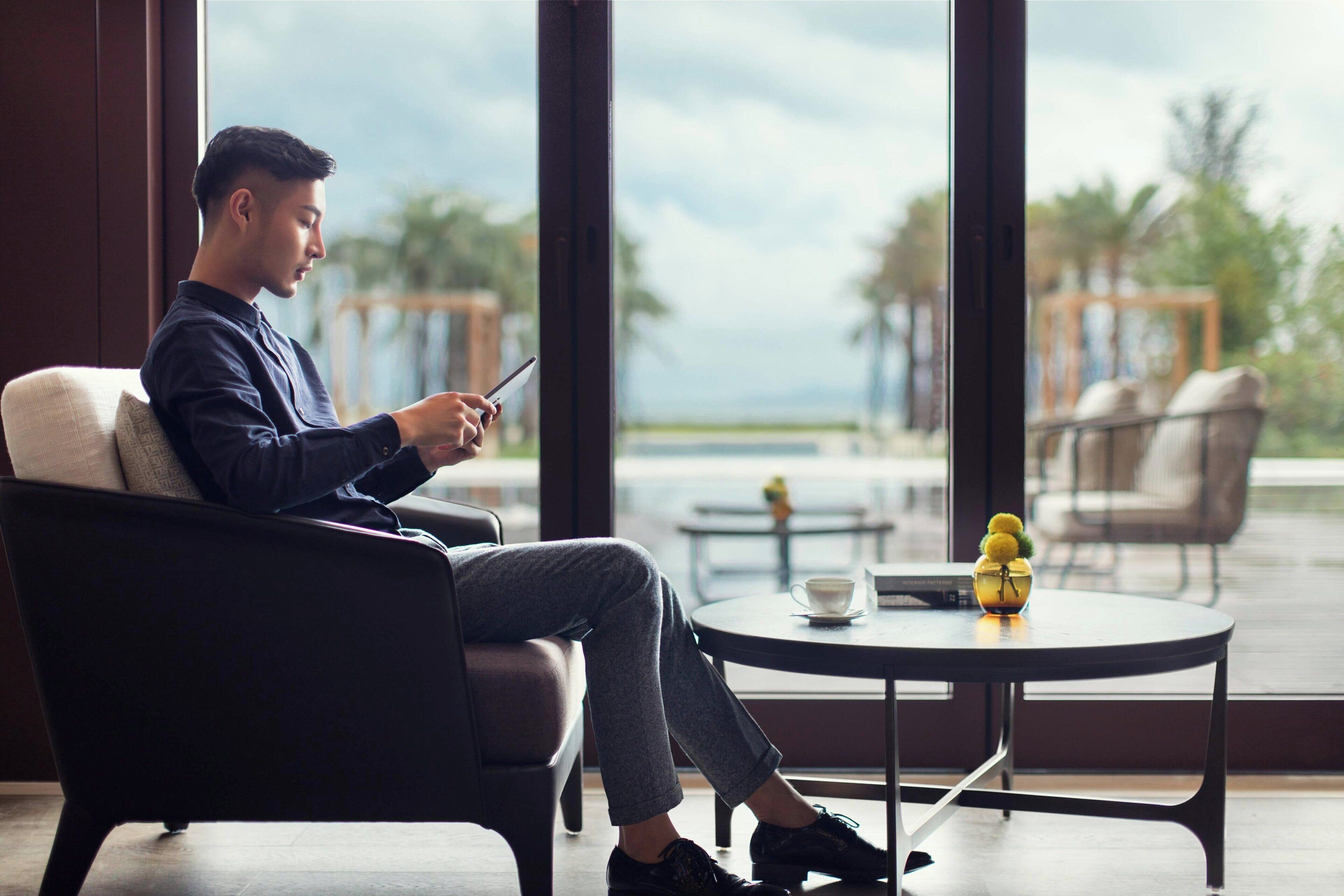 A man sitting on a chair by a window, using a smartphone. There is a table with a cup and a decorative item.