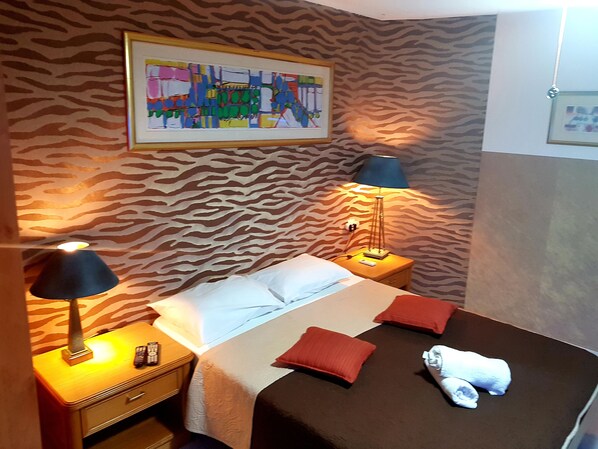Une chambre d’hôtel avec un mur à motifs zébrés, un lit avec des oreillers blancs et rouges, une serviette blanche posée sur le lit et un tableau abstrait encadré au-dessus du lit.
