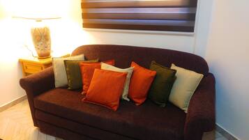 A dark brown sofa with multiple cushions in various colors, a small wooden side table with a lamp, and a wall-mounted light fixture.
