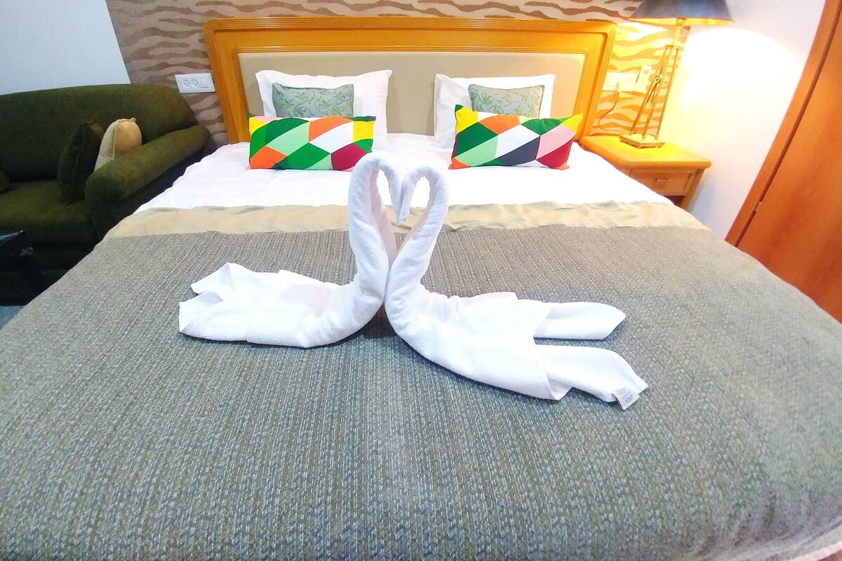 Una cama bien hecha con una escultura de toalla, un sillón verde y cojines estampados.