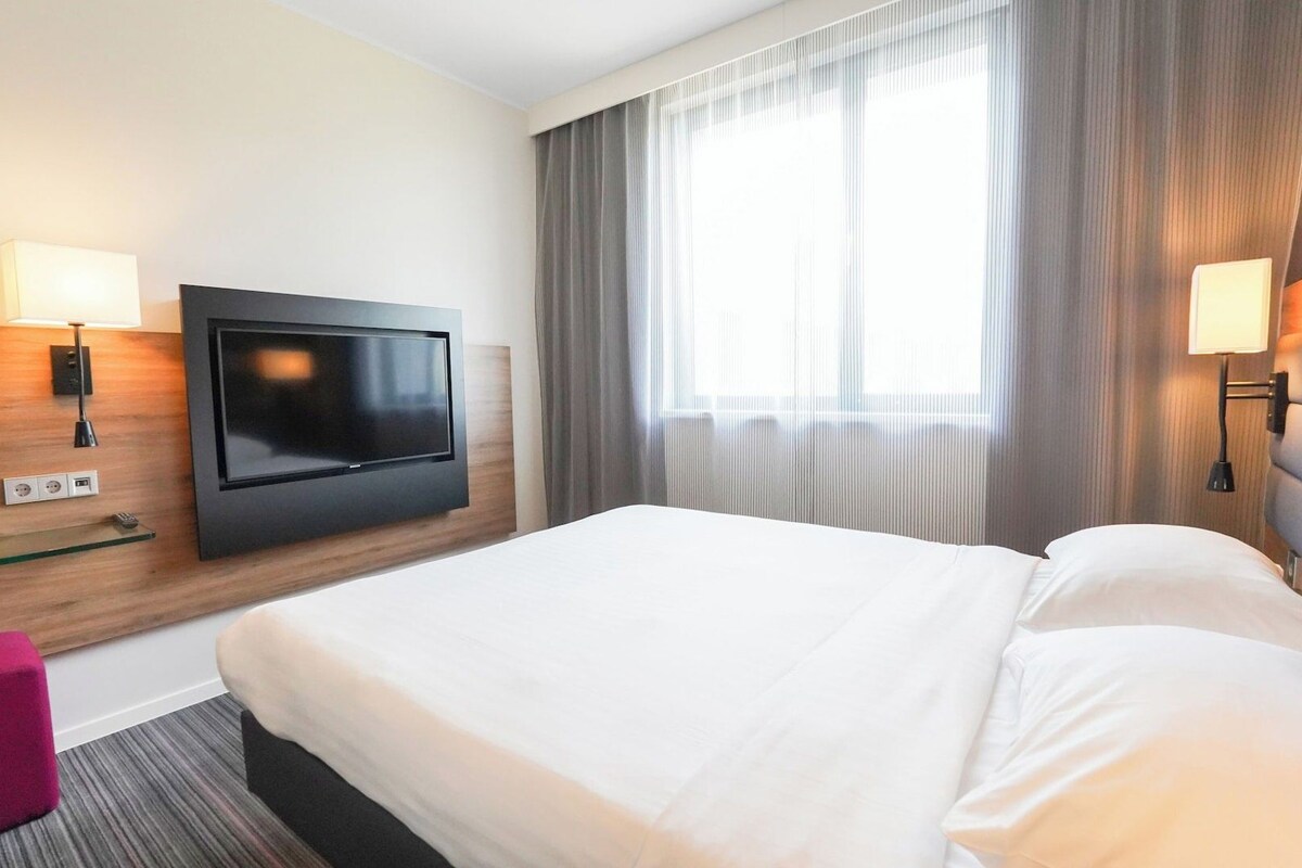 Habitación de hotel con televisor de pantalla plana montado en la pared, cama con ropa blanca, un pequeño taburete rosa y una ventana con cortinas.