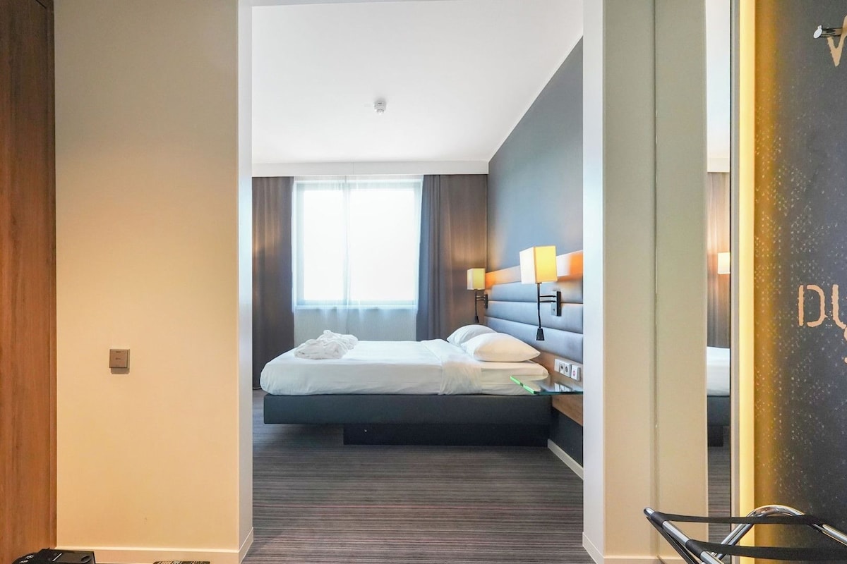 Una habitación de hotel con una cama, lámparas de noche, un ventanal con cortinas y una puerta de madera.