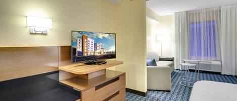 Habitación de hotel con televisión de pantalla plana sobre un soporte de madera, un escritorio con cajones, una silla acolchada, una mesita redonda pequeña y una cama grande.