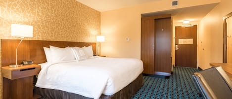Een hotelkamer met een groot bed, een nachtkastje met een lamp, een houten hoofdeinde, een kamerbreed tapijt met een patroon, en een deur die naar een andere kamer leidt.