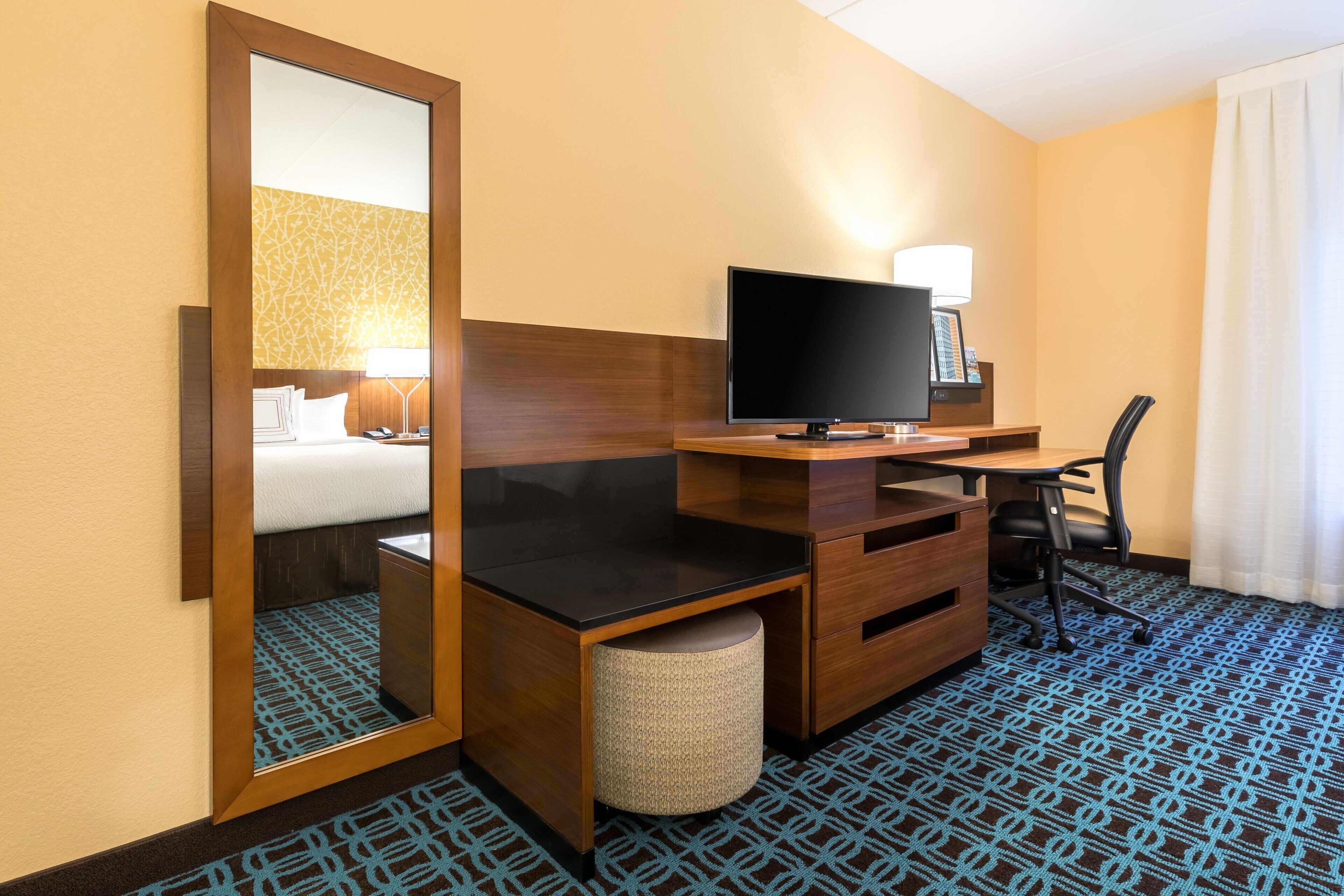 A hotel room with a bed, a desk with a computer, a chair, a lamp, and a mirror.