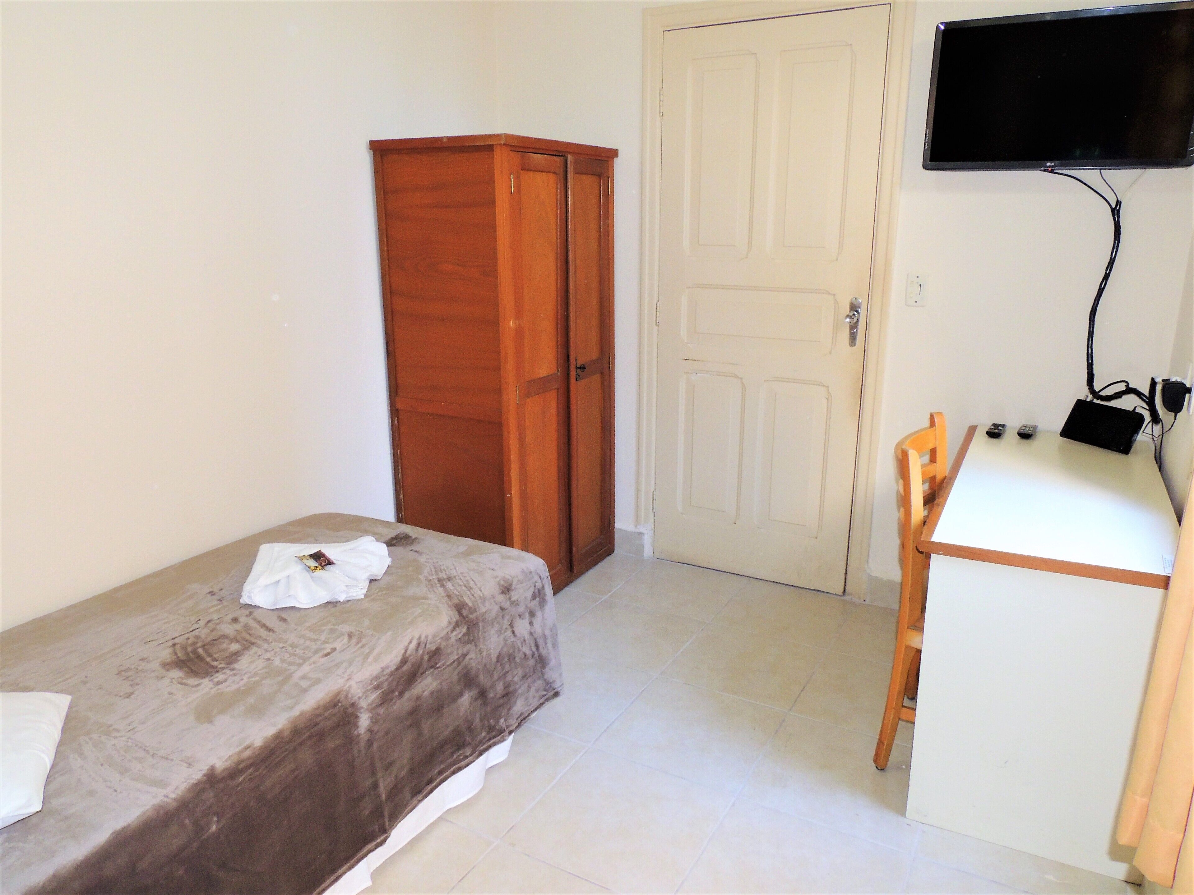 Una habitación pequeña con una cama, un armario de madera, un escritorio con una silla y un televisor en la pared.