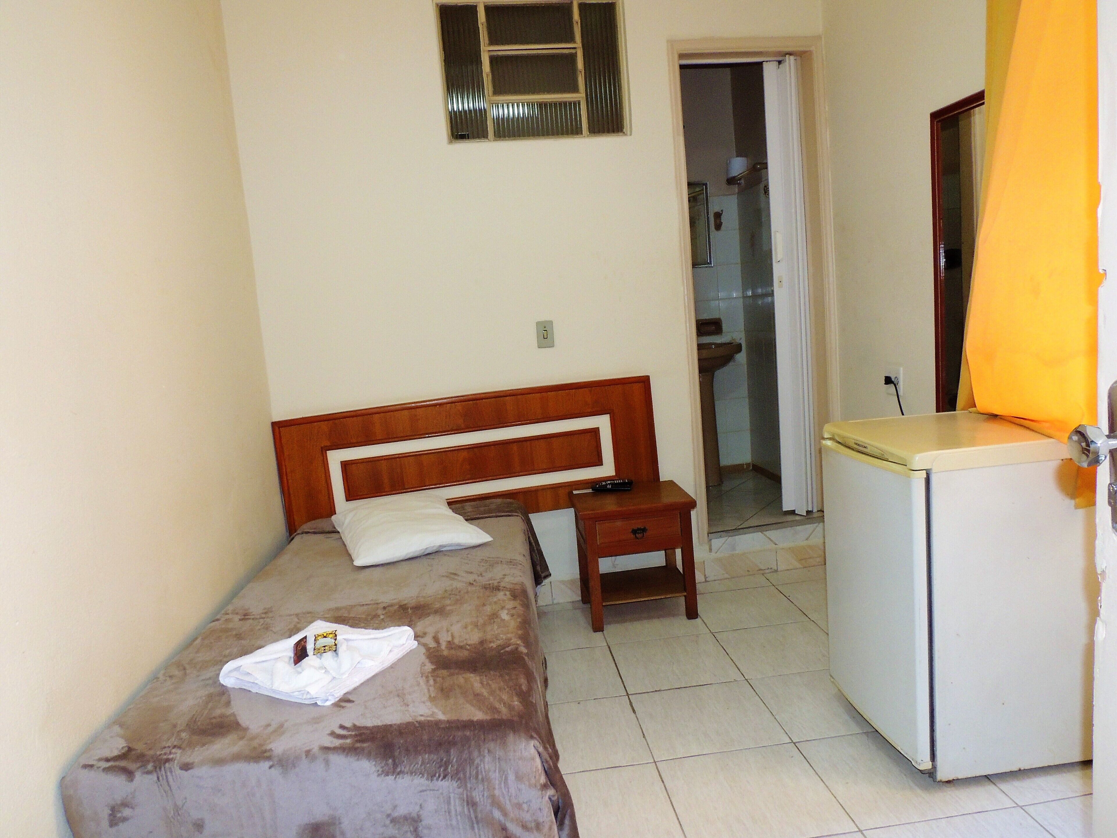 A small, single-bed room with a wooden headboard, a bedside table, a small cabinet, and a bathroom visible through an open door.