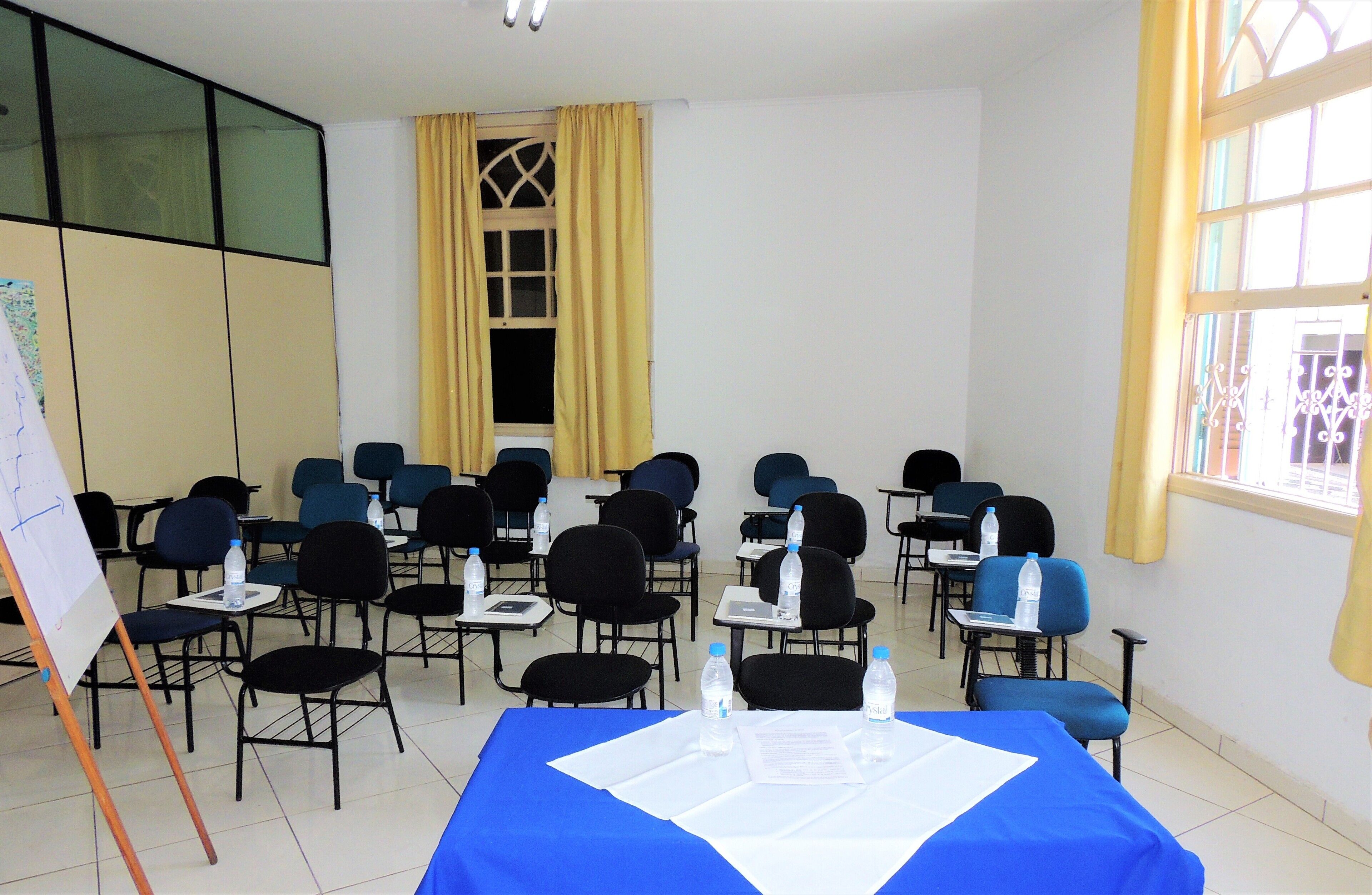 Uma sala de conferências com fileiras de cadeiras, uma mesa azul com papéis e garrafas de água, e uma janela grande com cortinas amarelas.
