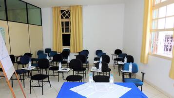 Uma sala de conferências com fileiras de cadeiras, uma mesa azul com papéis e garrafas de água, e uma janela grande com cortinas amarelas.