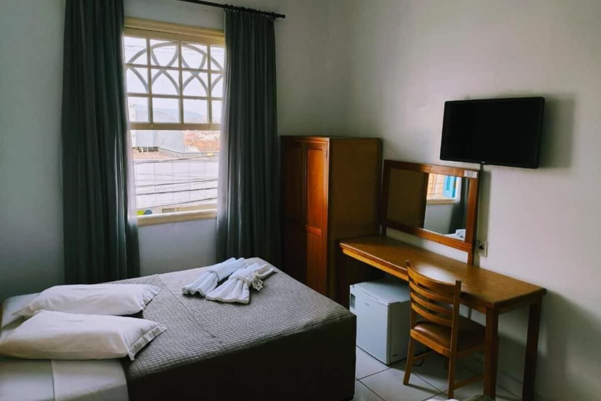 Una habitación de hotel con cama, escritorio, televisor y ventana con cortinas.