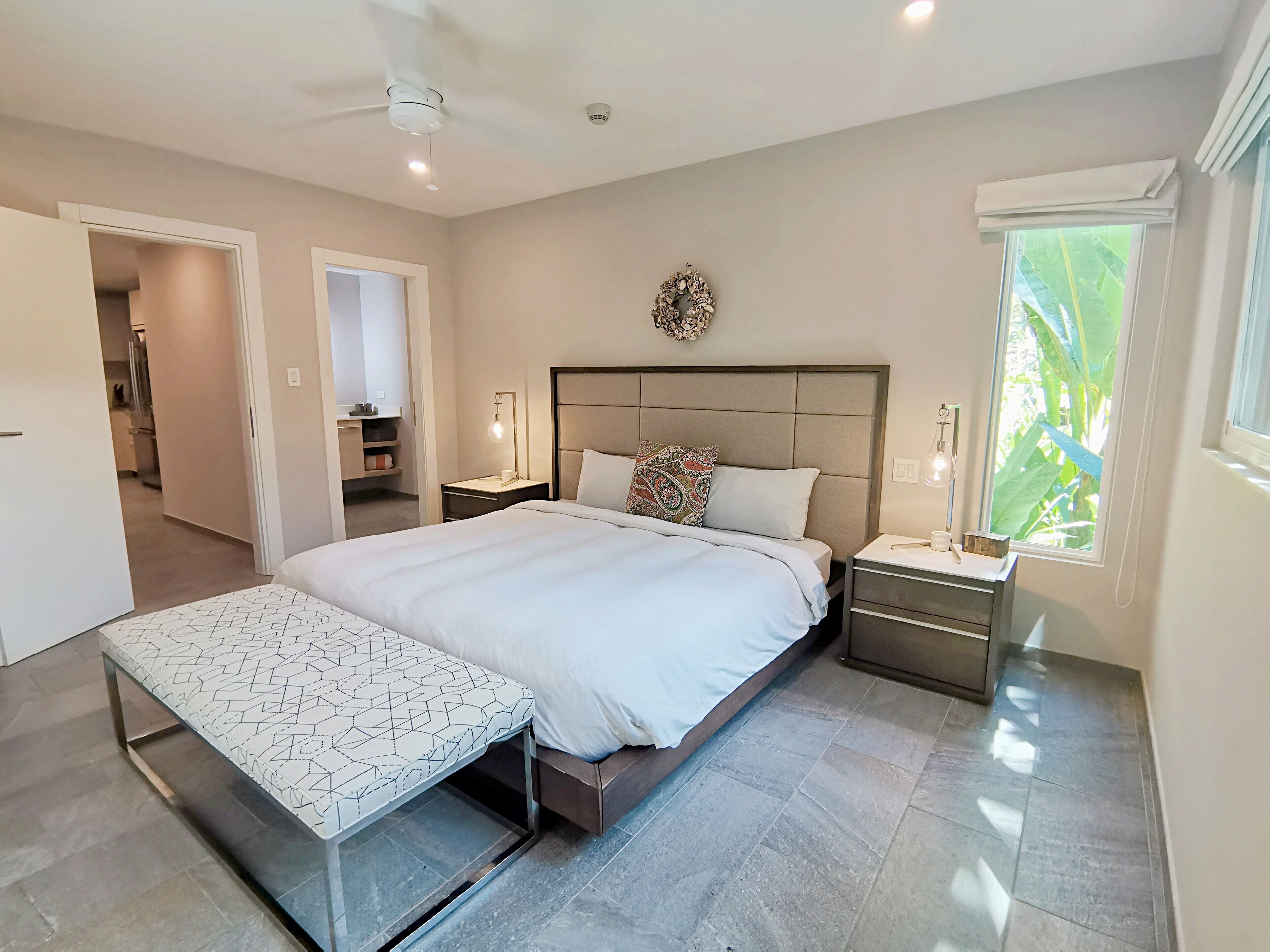 A modern bedroom with a large bed, bedside tables, a bench, and a view of greenery through the window.