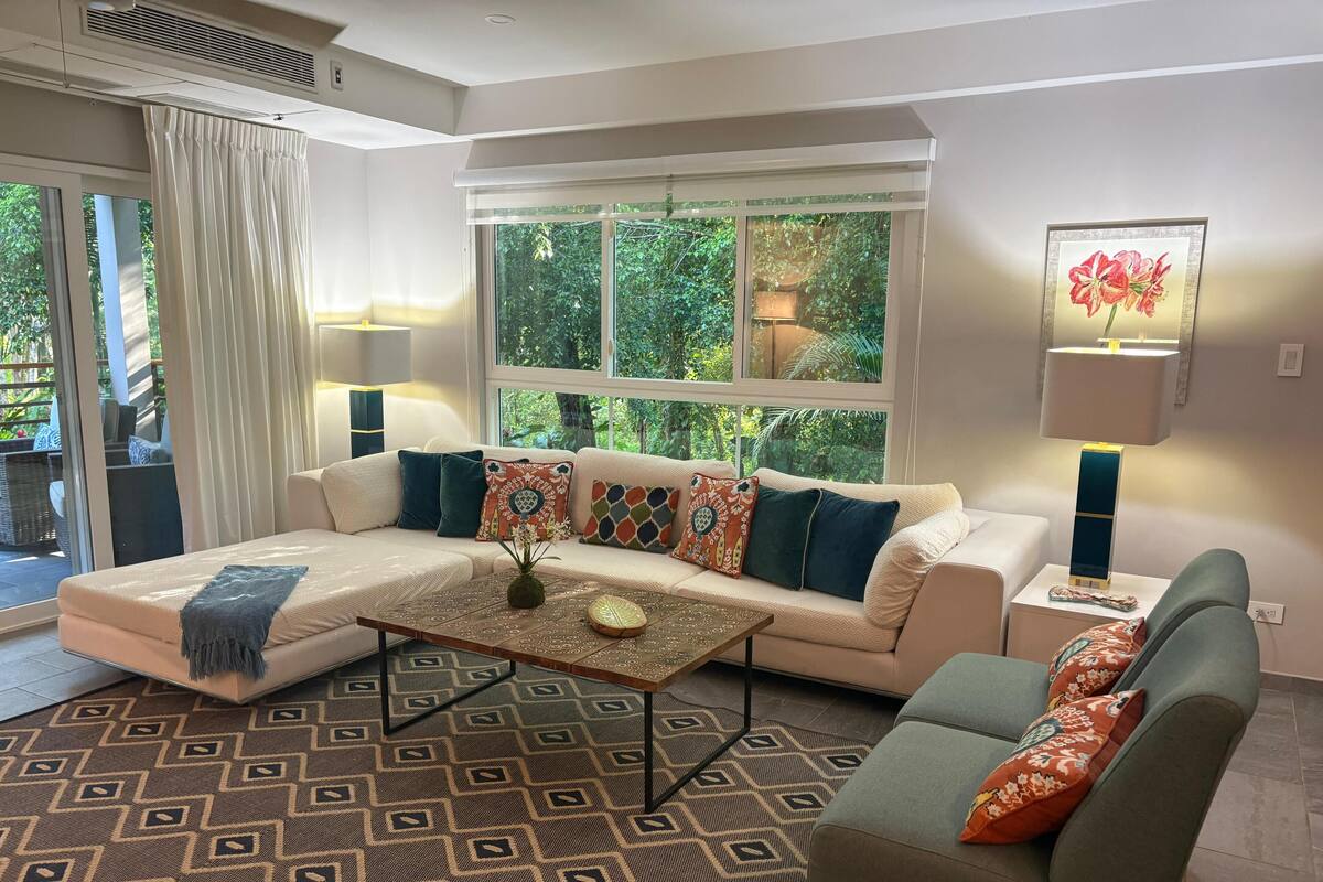 A modern living room with a sectional sofa, patterned rug, and a view of greenery through large windows.