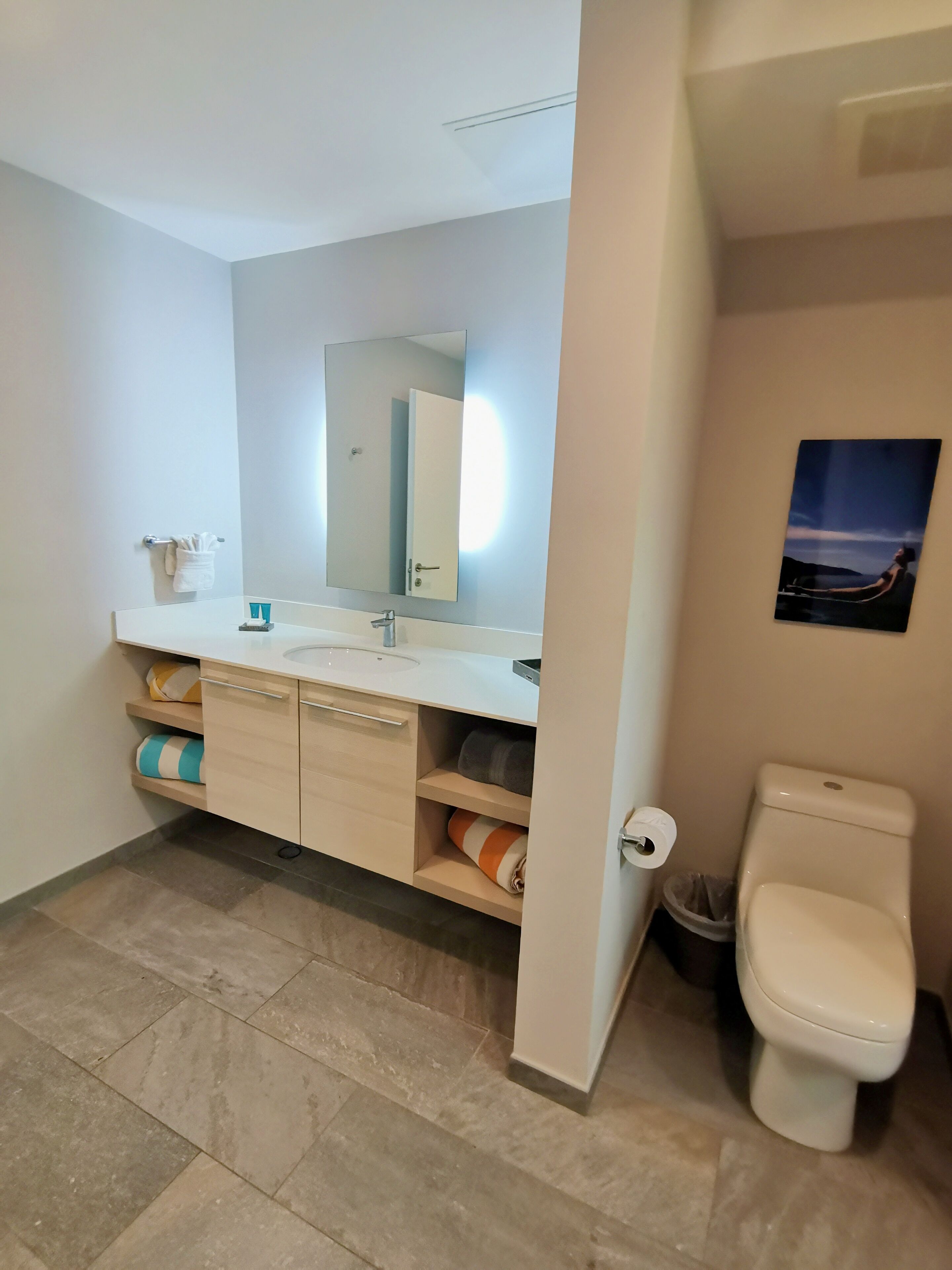 A bathroom with a white sink, mirror, and toilet. There are towels and a trash bin visible.