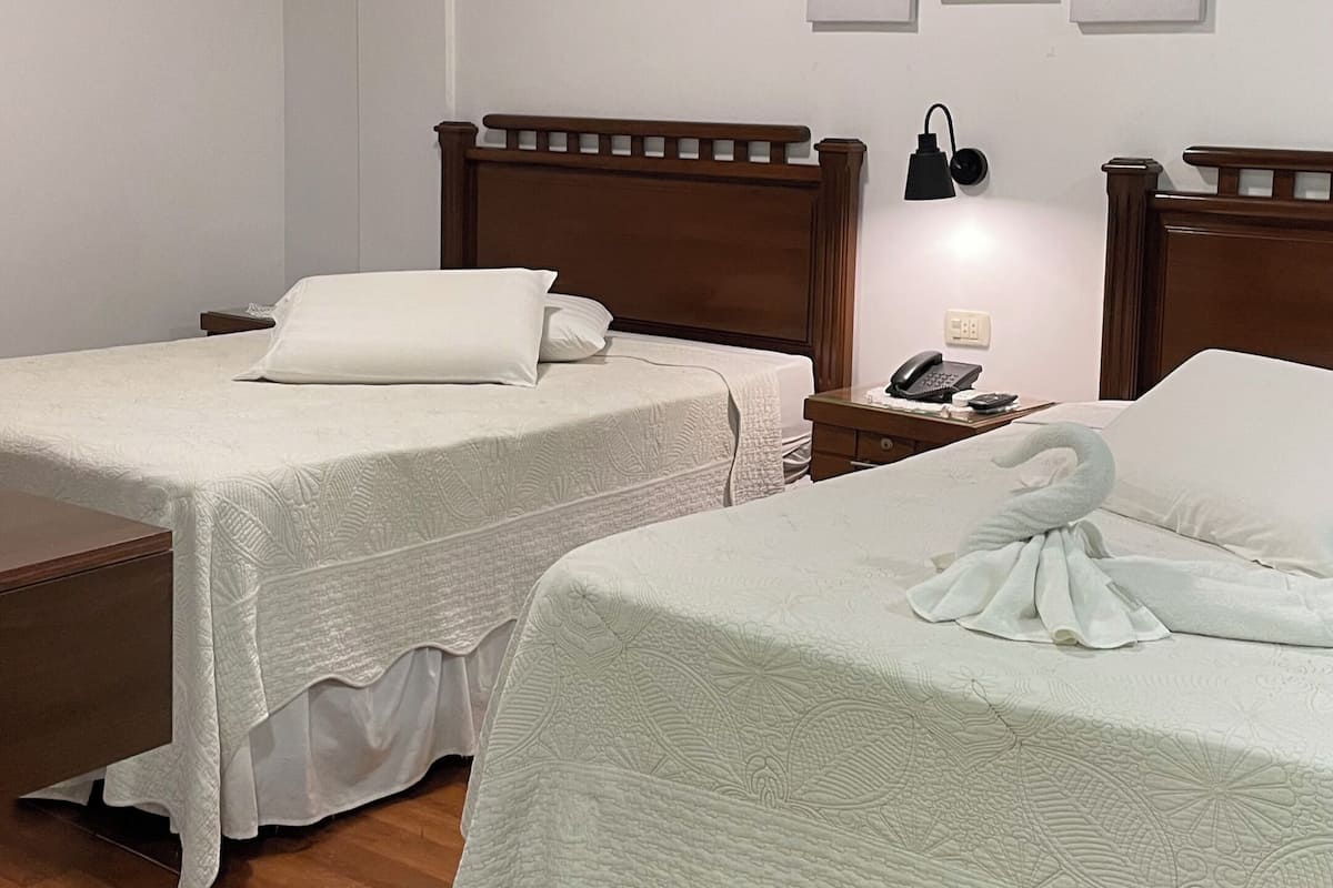 Habitación de hotel con dos camas, cabeceros de madera, una mesita de noche con lámpara y un teléfono en la pared.