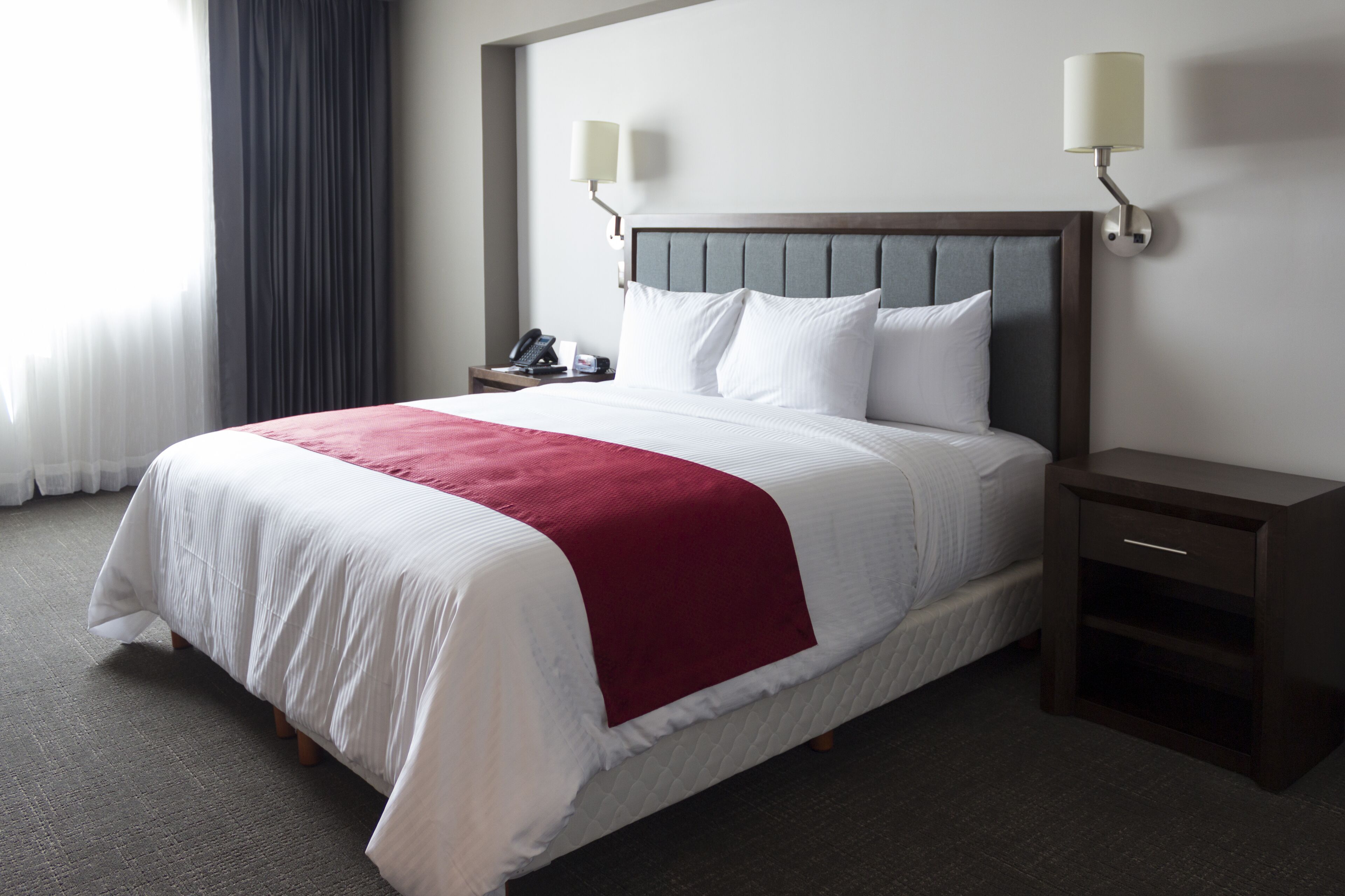A hotel room with a neatly made bed, a bedside table with a lamp, and a nightstand with a phone.