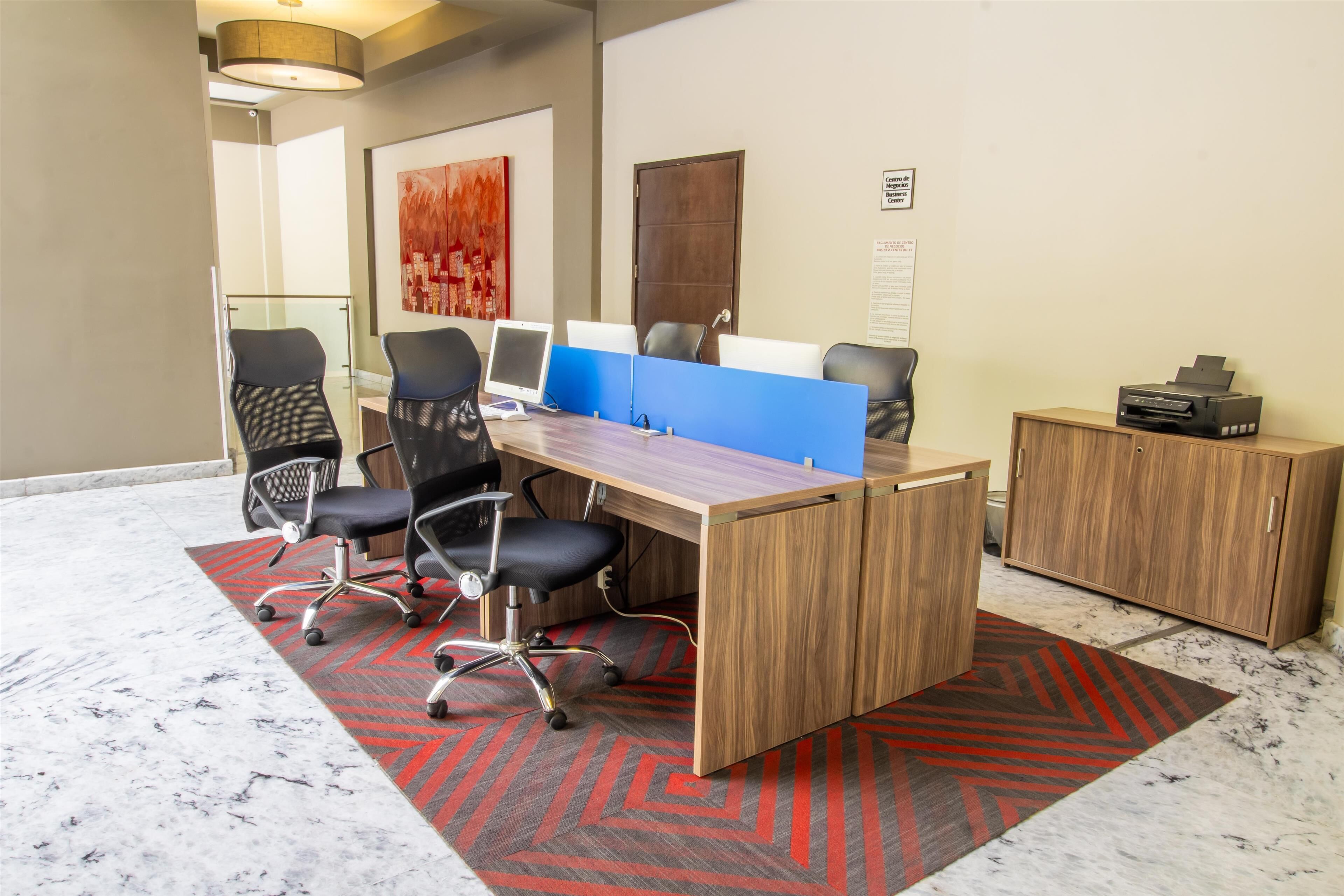 A modern office space with a long wooden desk, black office chairs, a glass partition, and a printer on a cabinet.
