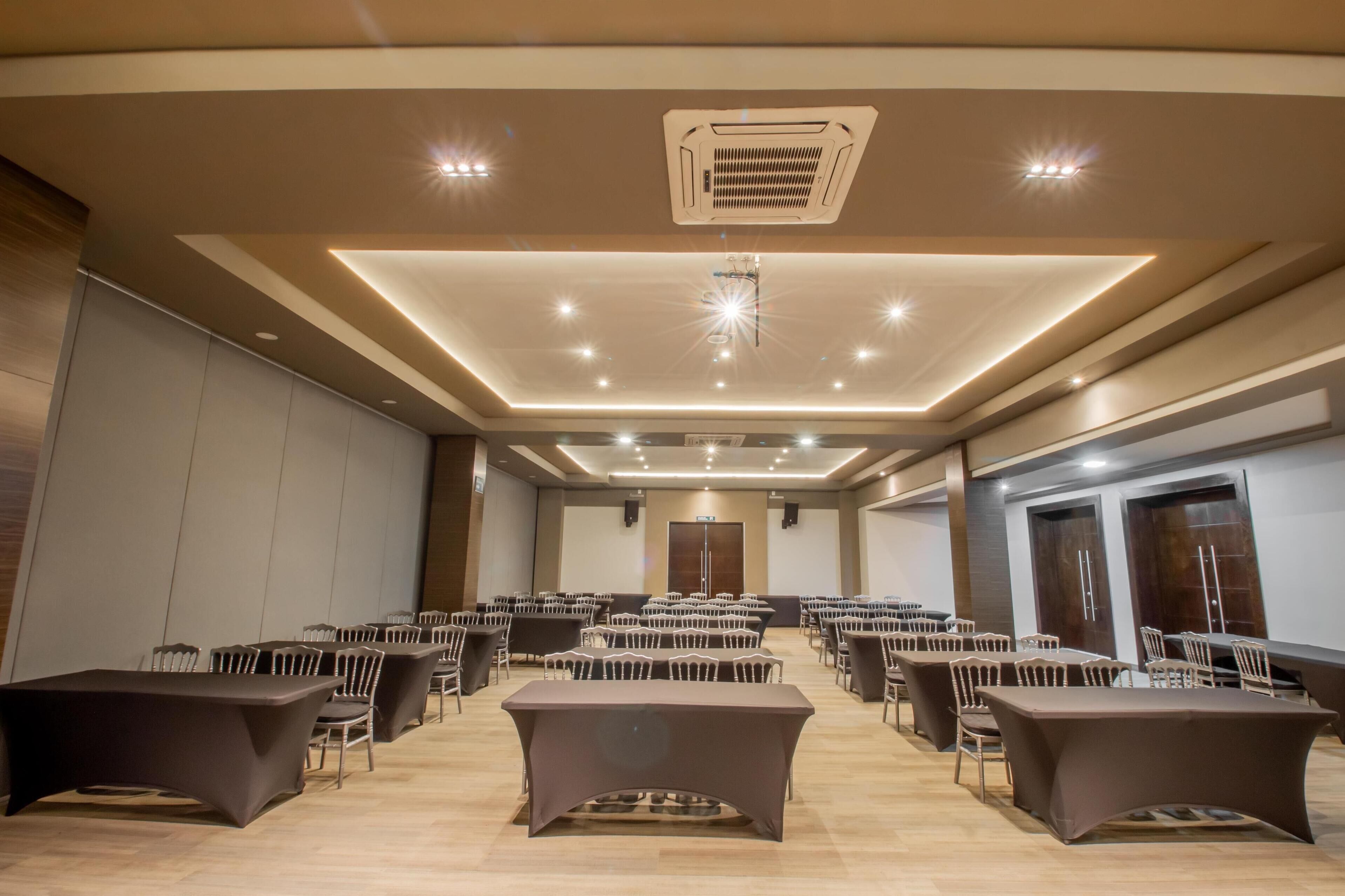 Un salón de conferencias con varias mesas y sillas, iluminación empotrada y un respiradero en el techo.