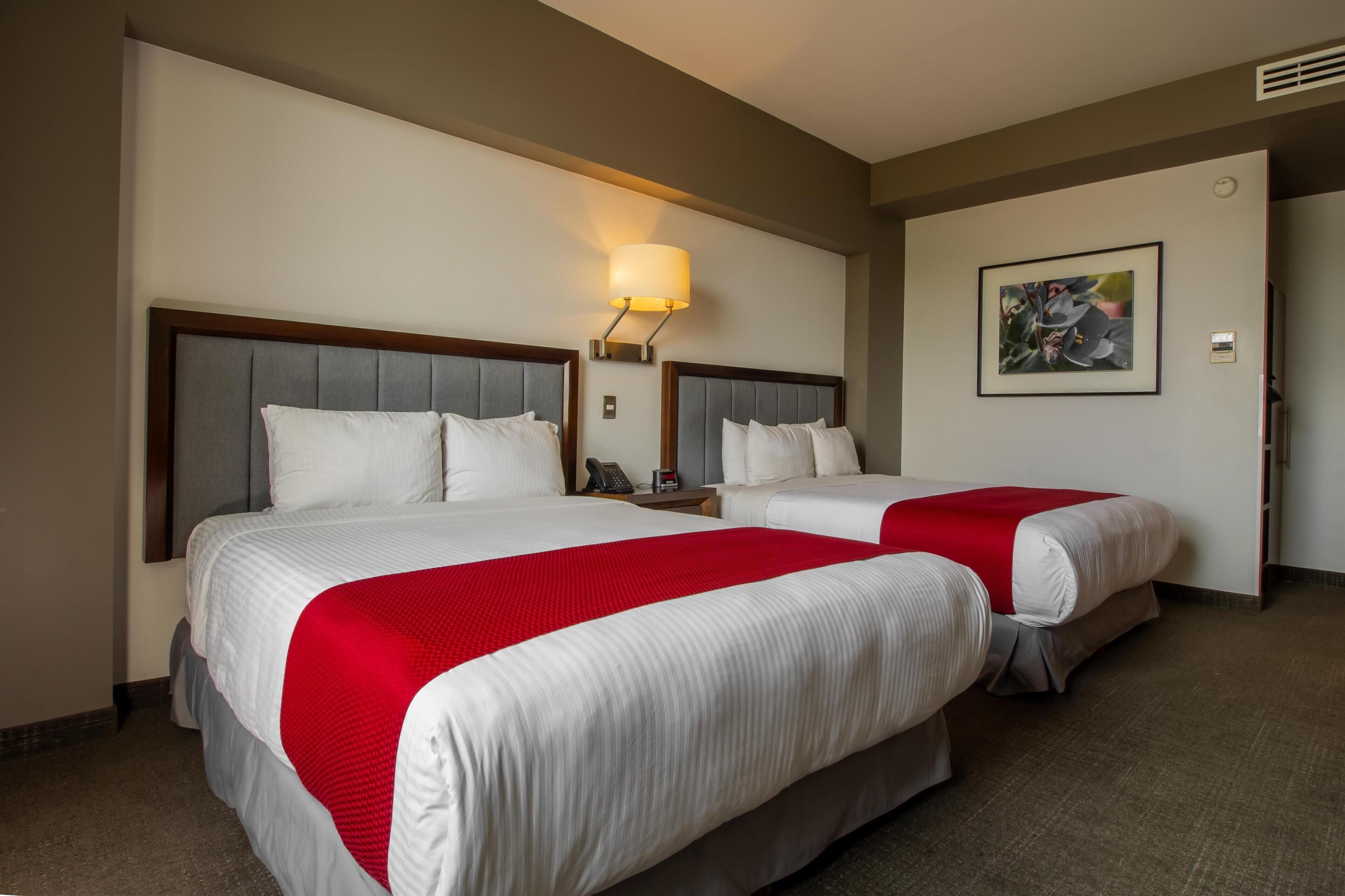 A hotel room with two beds, a bedside table with a lamp, a framed picture on the wall, and a door.