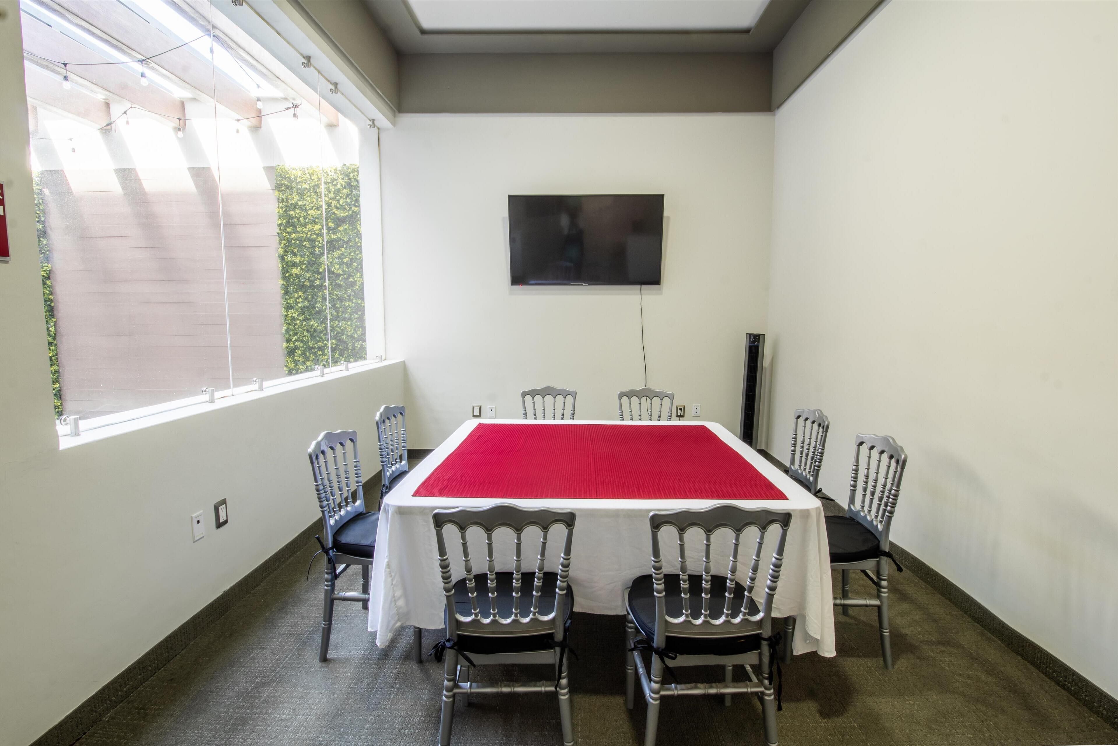 Un salón de conferencias con una gran mesa, seis sillas, un televisor en la pared y un ventanal que proporciona luz natural.