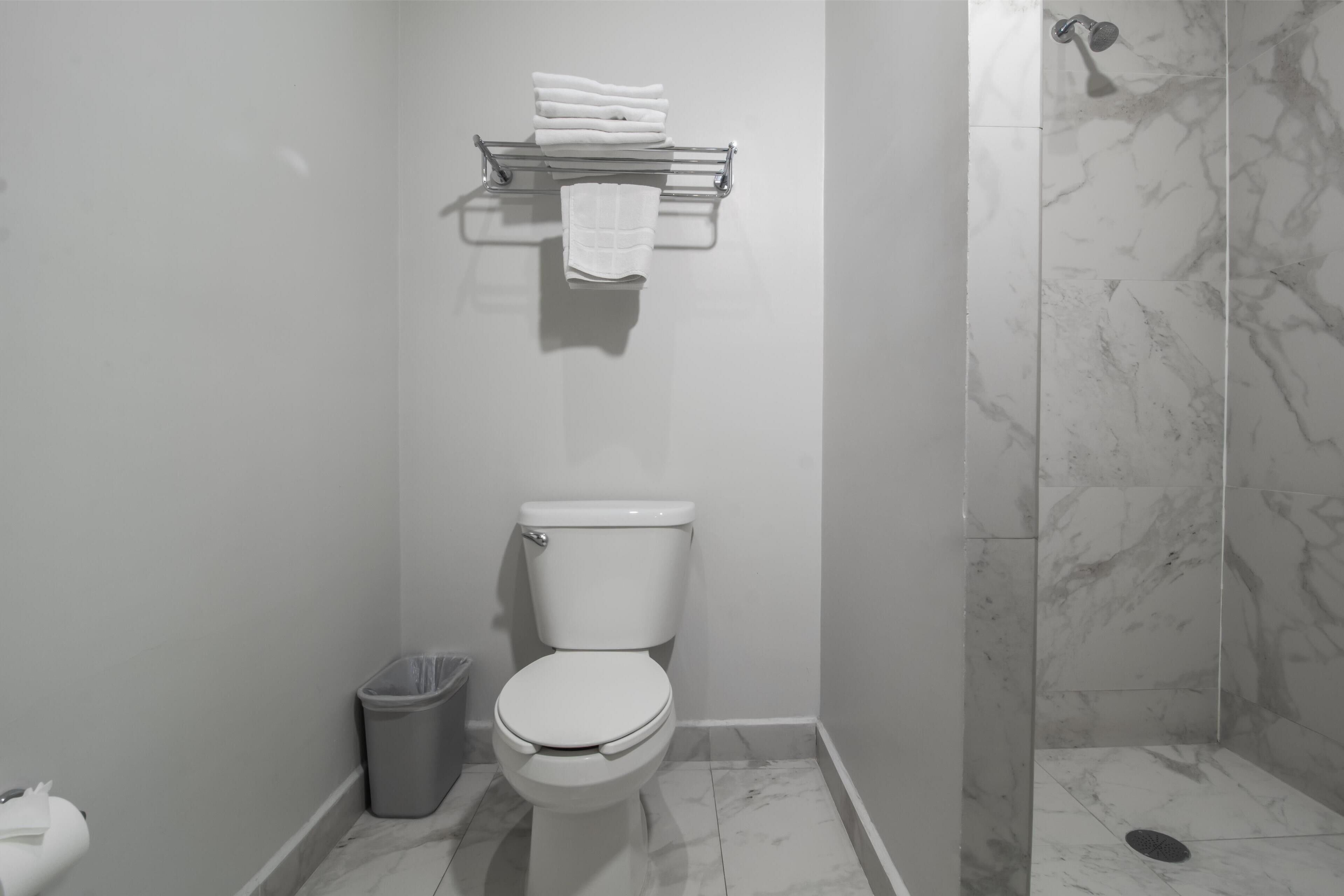 A bathroom with a toilet, a trash bin, a towel rack with folded towels, and a shower area with a drain.