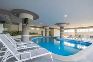 An indoor swimming pool with white lounge chairs, a camera on a pillar, and a ceiling with recessed lighting.