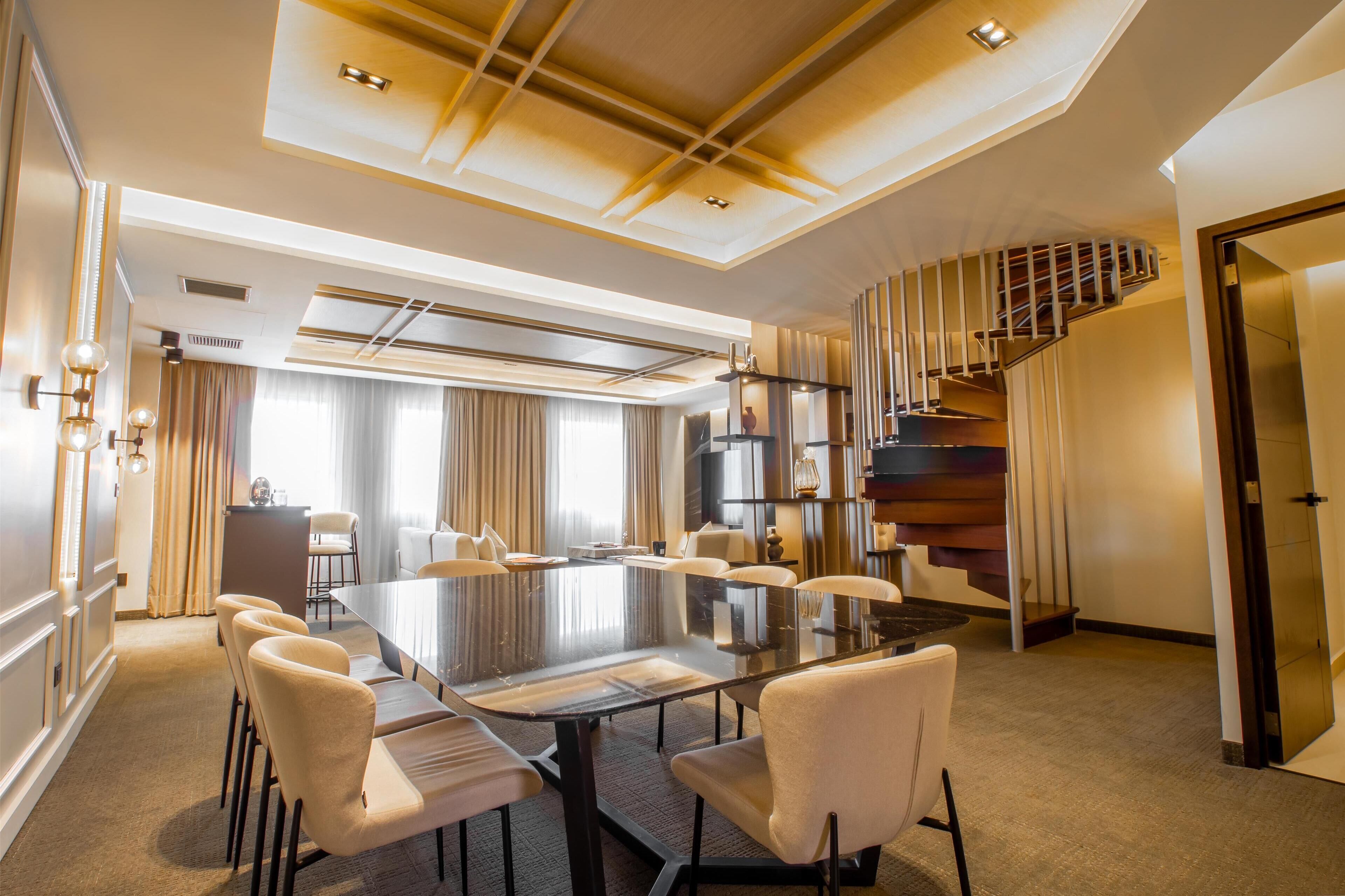 A spacious conference room with a large table and comfortable chairs, a modern staircase, and a well-lit ceiling with recessed lighting.