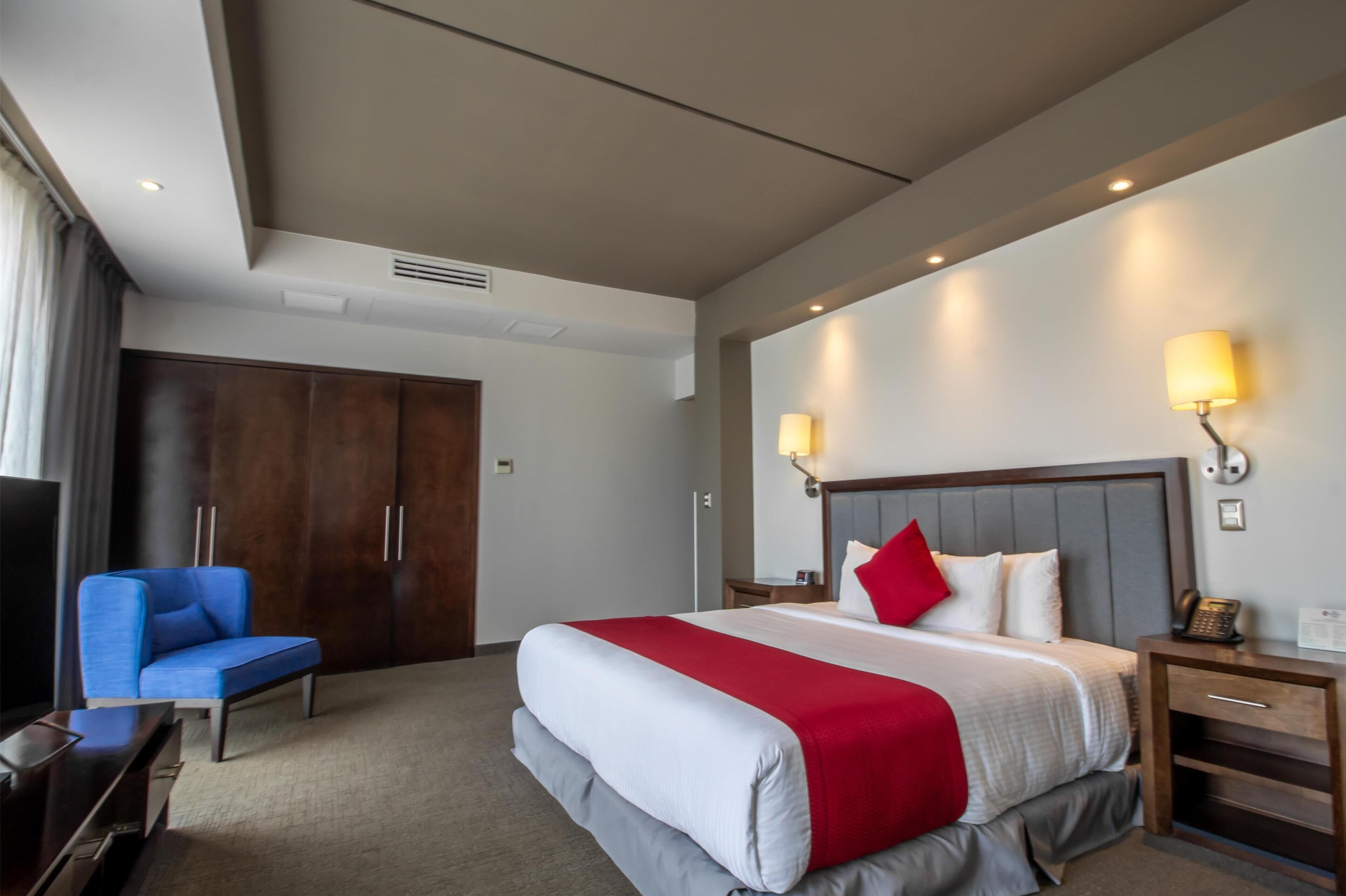Una habitación de hotel con una cama grande, una silla azul, un armario de madera y dos lámparas de noche.