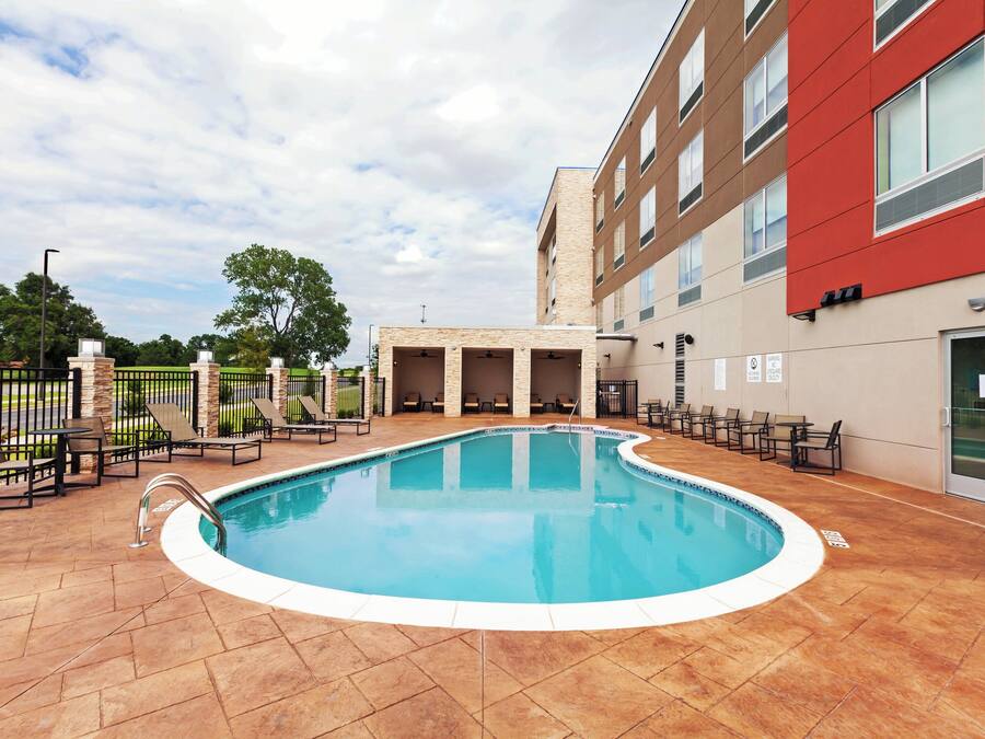 A swimming pool with a tiled deck, surrounded by lounge chairs and a building with multiple windows.