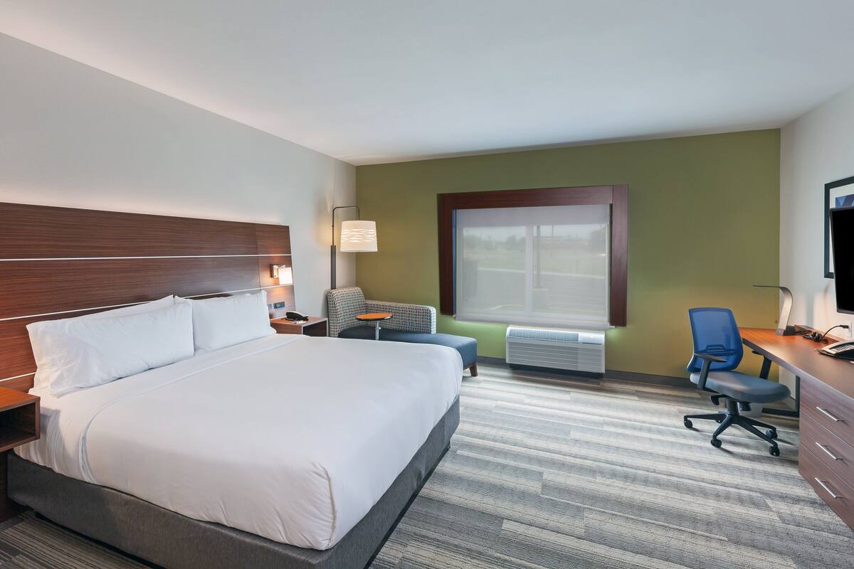 Une chambre d’hôtel avec un grand lit, un bureau avec une chaise, une petite table et une fenêtre avec des stores.