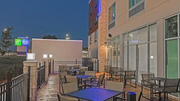 An outdoor patio area with tables and chairs, a stone wall, and a building with large windows.