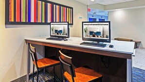 Un comptoir d’accueil d’hôtel avec deux écrans d’ordinateur, un panneau indiquant le centre d’affaires et une œuvre d’art colorée à rayures accrochée au mur.