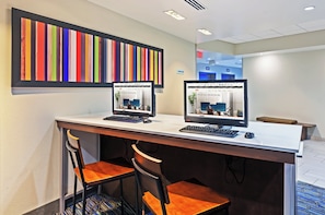 Un comptoir d’accueil d’hôtel avec deux écrans d’ordinateur, un panneau indiquant le centre d’affaires et une œuvre d’art colorée à rayures accrochée au mur.