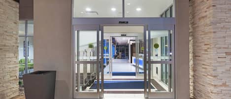 Ein Hotel Eingang mit automatischer Schiebetür, einer Rezeption und einer Wartezone mit Stühlen und einem blauen Teppich.