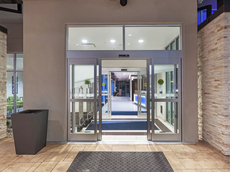 Entrada de hotel con puertas correderas automáticas, una recepción y un área de espera con sillas y una alfombra azul.