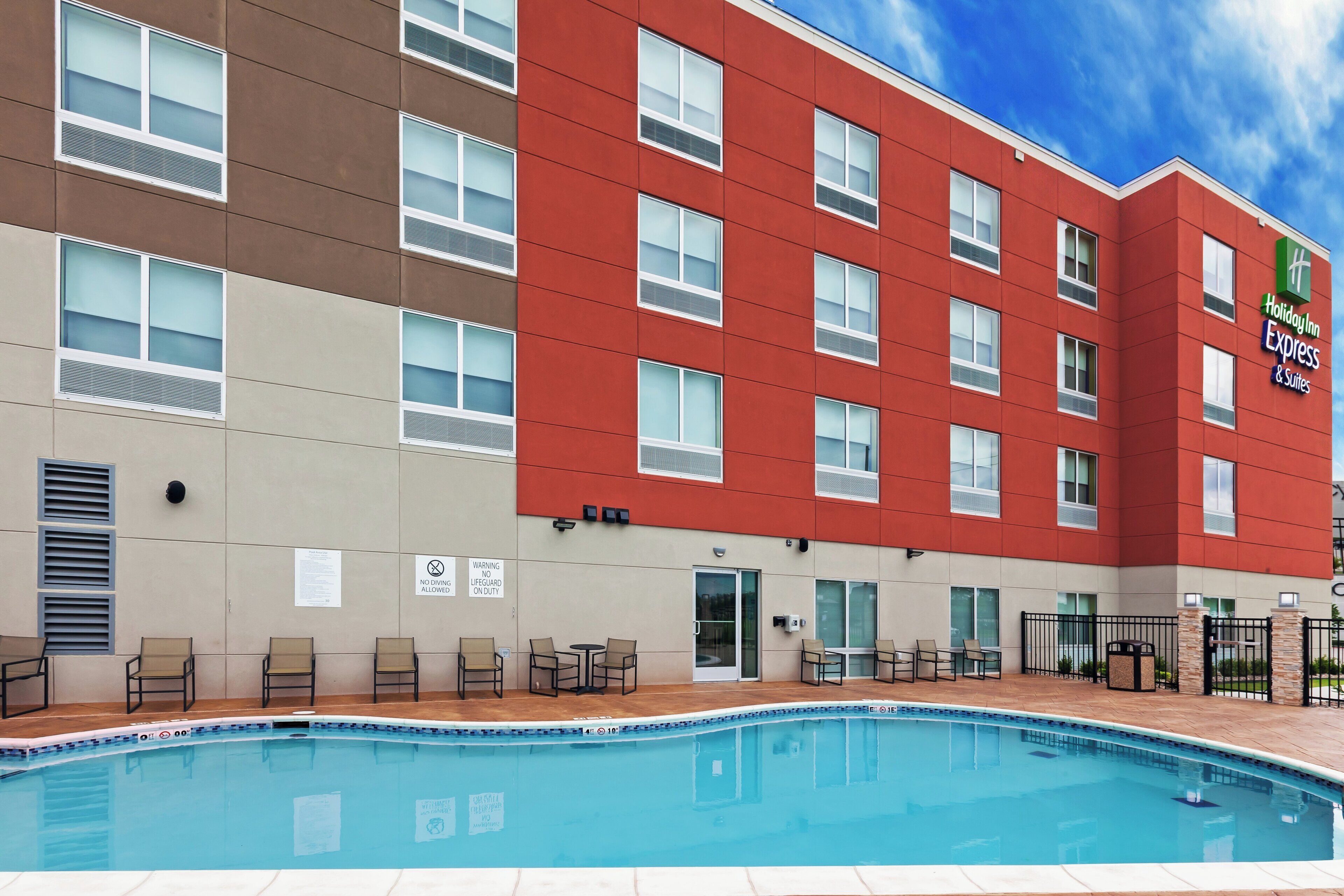 A Holiday Inn Express & Suites building with a swimming pool, outdoor seating, and a clear sky.