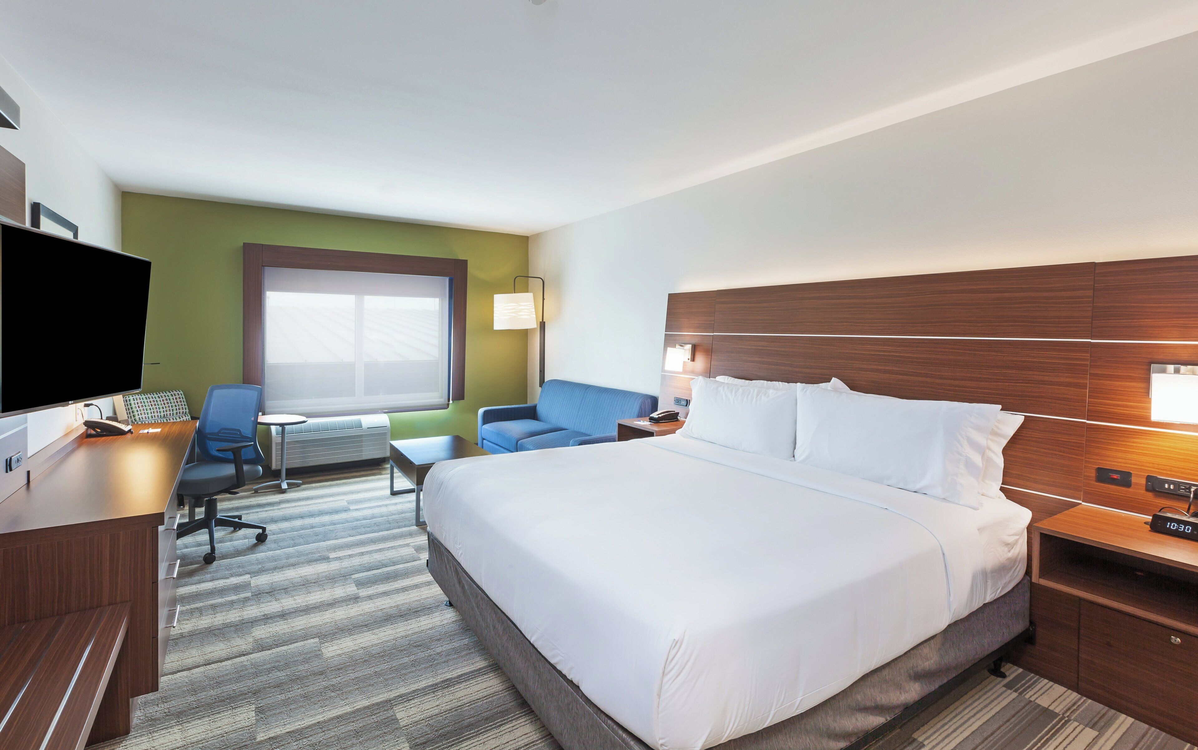A hotel room with a large bed, a desk with a computer, a chair, a sofa, and a TV.