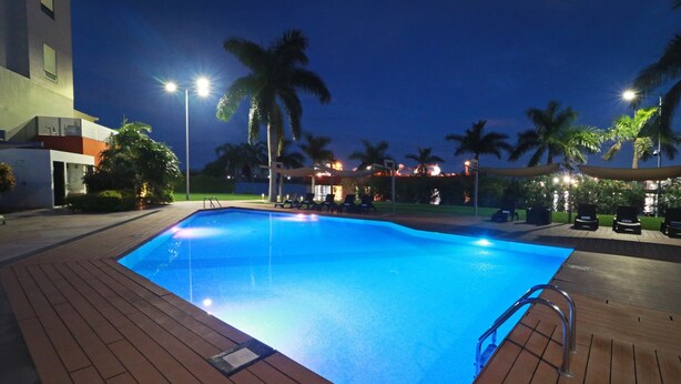 A rectangular swimming pool with a wooden deck, surrounded by palm trees and lounge chairs, with a building in the background.