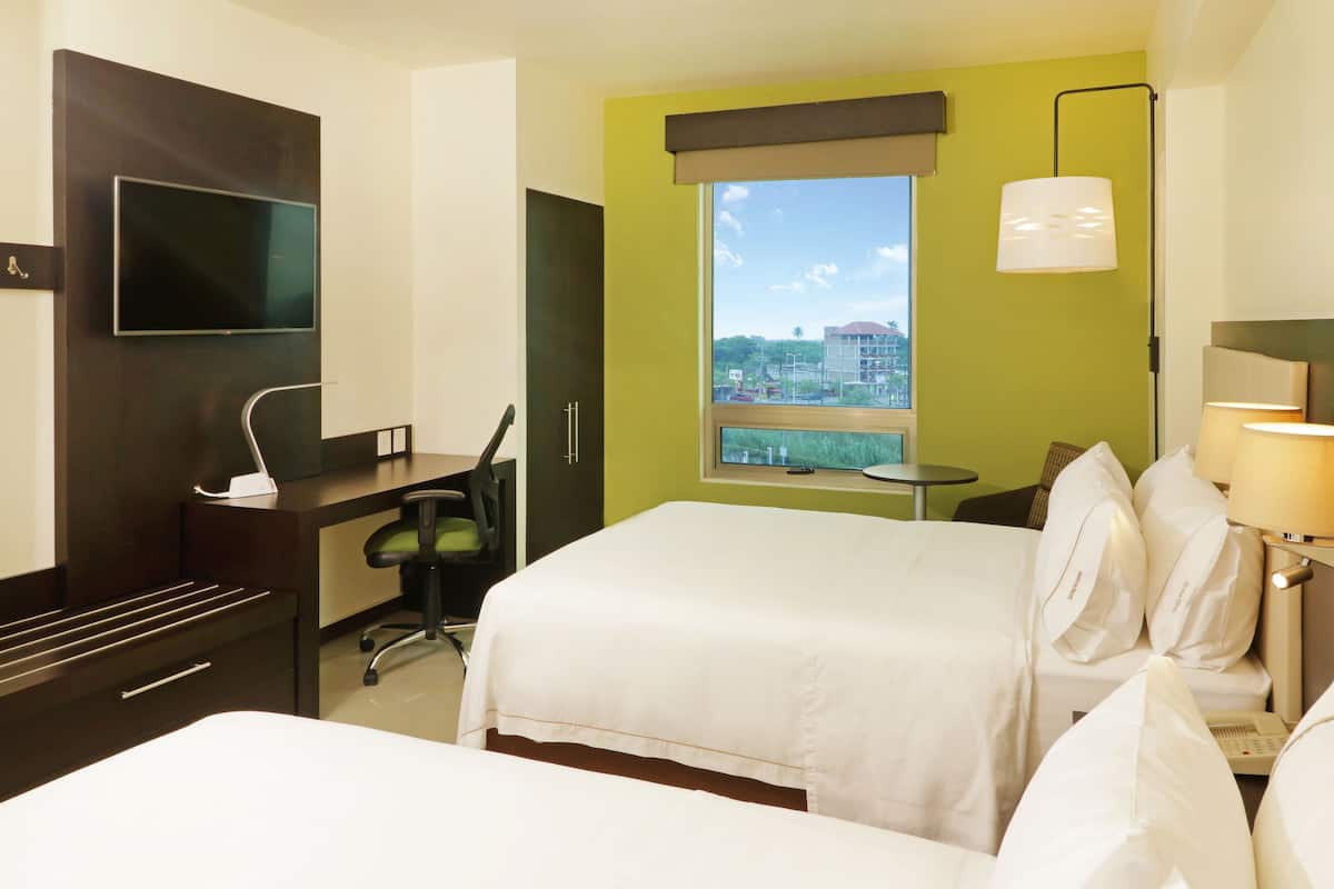 Quarto de hotel com cama, escrivaninha, cadeira, televisão e janela com vista para prédios.