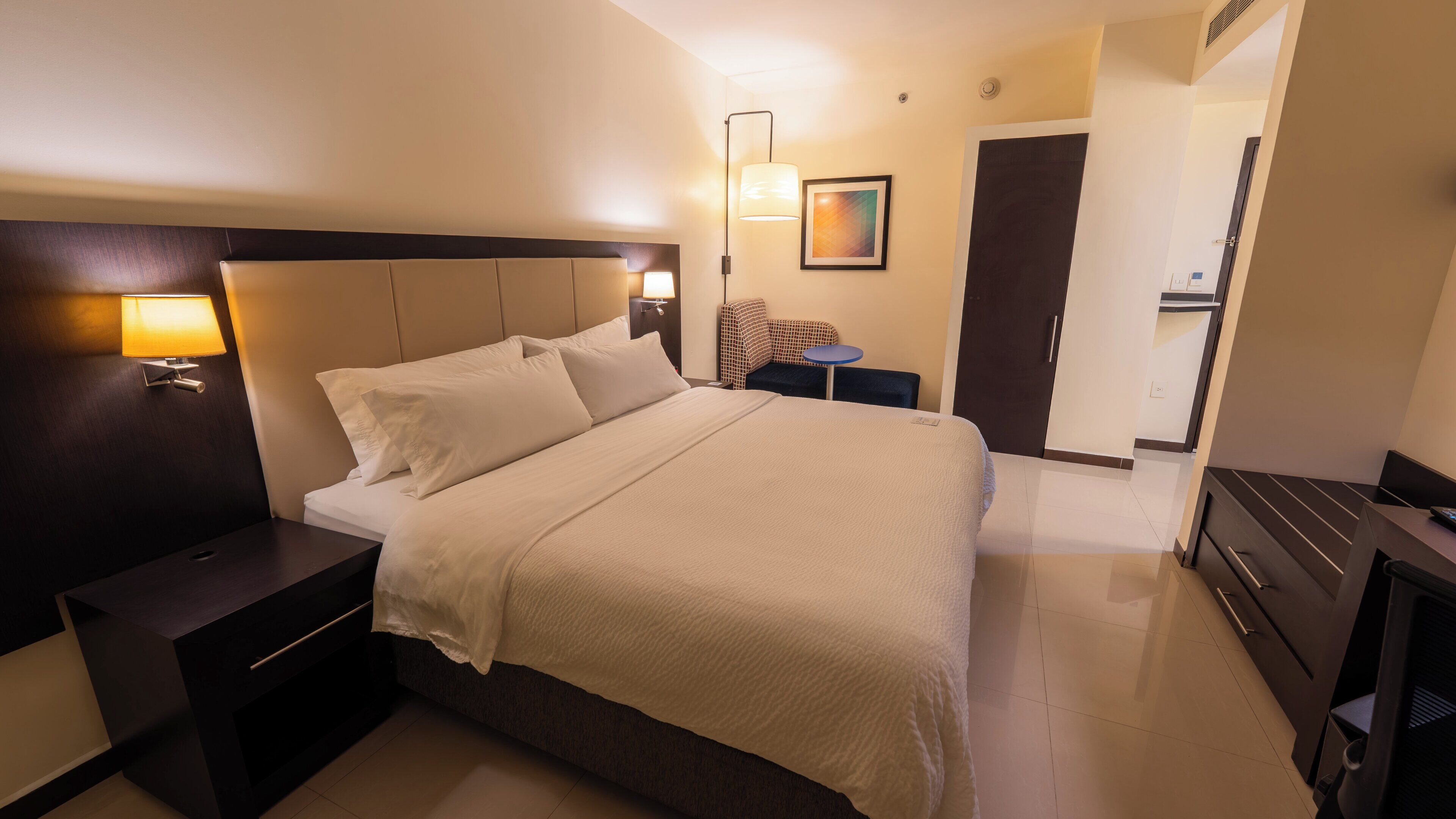 Habitación de hotel con cama, mesitas de noche, escritorio, silla, lámpara y puerta.